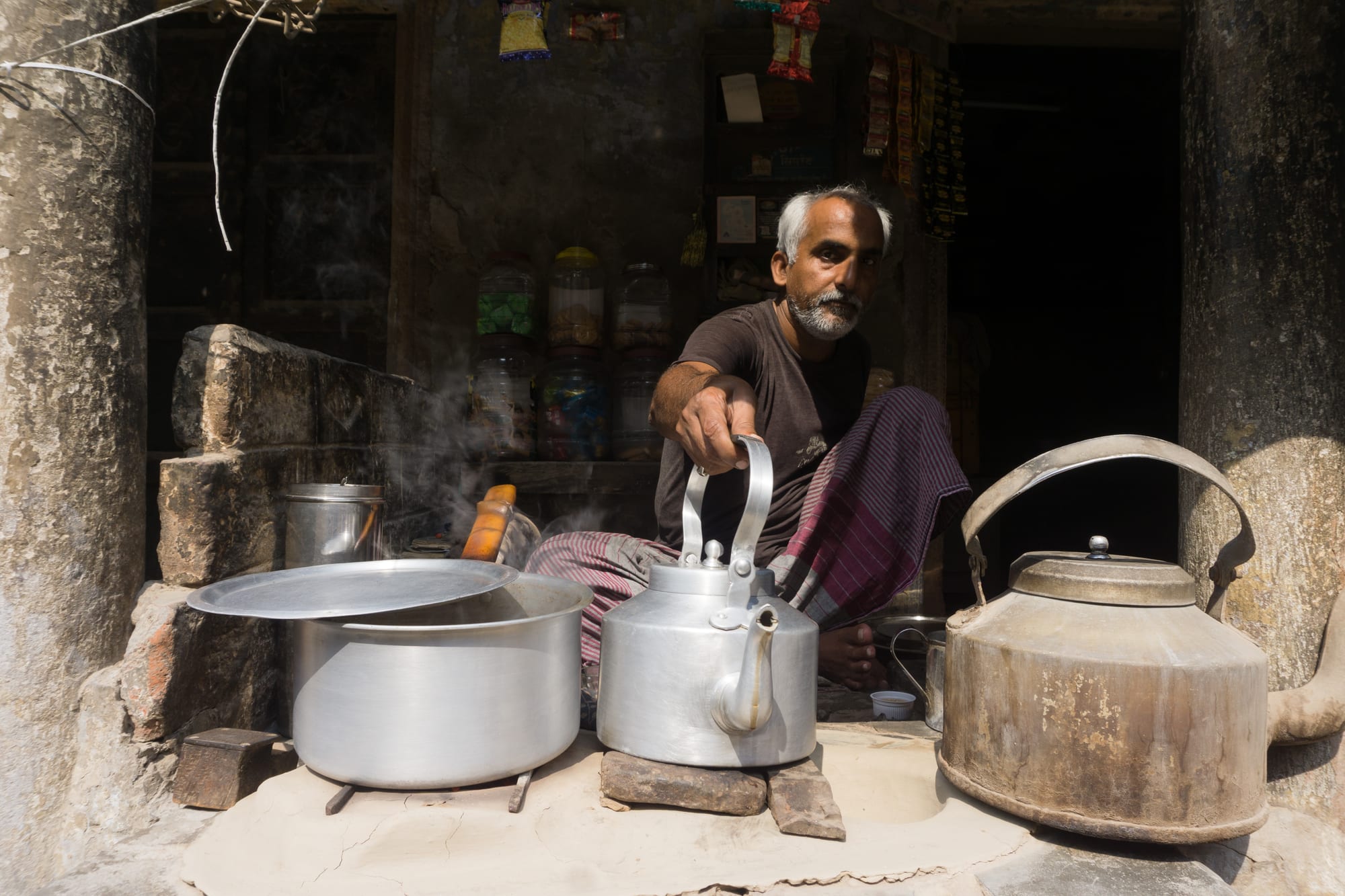 A tea seller sitting beside large kettles of boiling chai at a small roadside stall