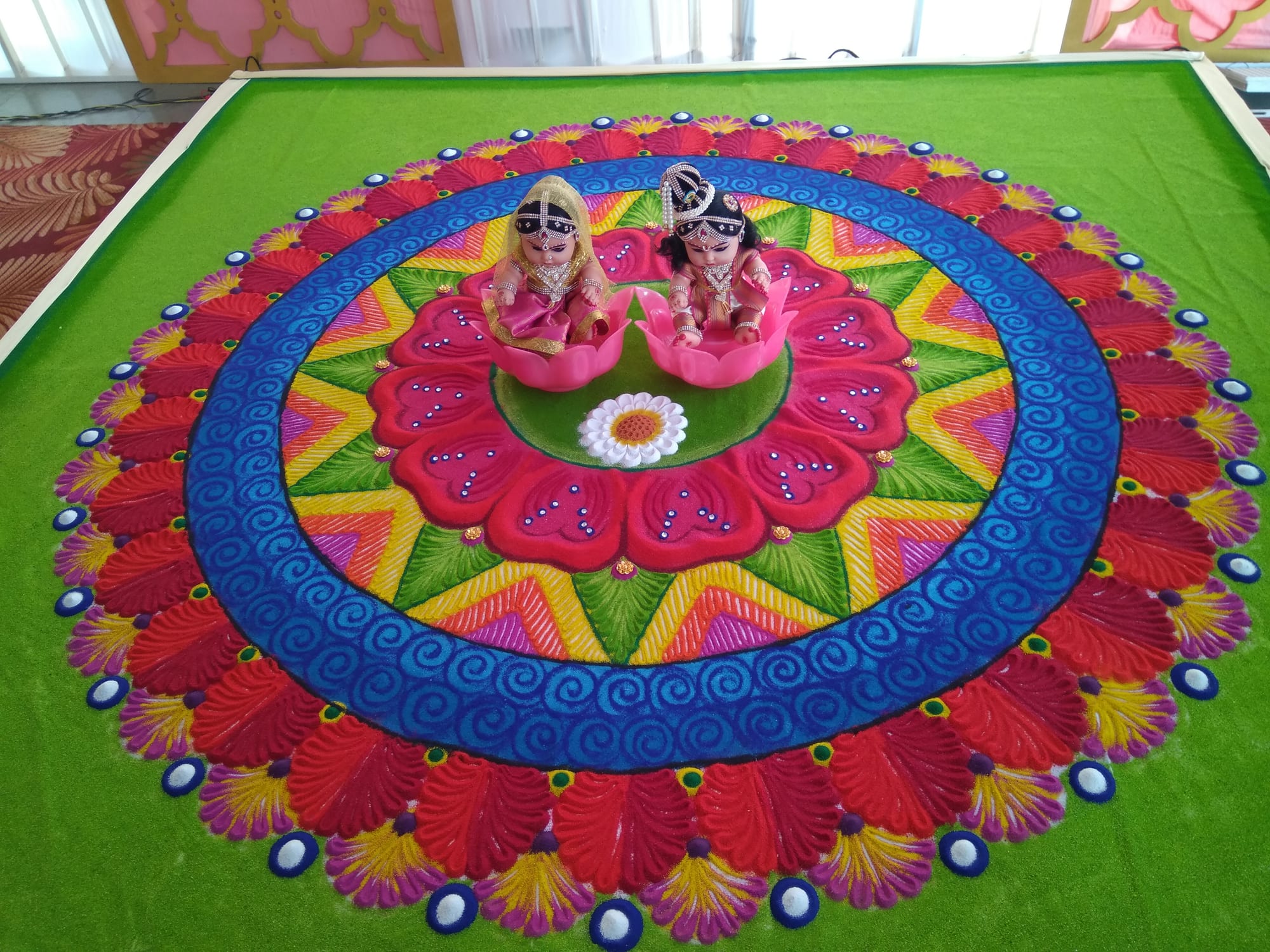 A large circular rangoli design in bright red, blue, yellow and pink coloured powder on a green base, with two small bride and groom dolls seated on pink lotus shapes at its centre