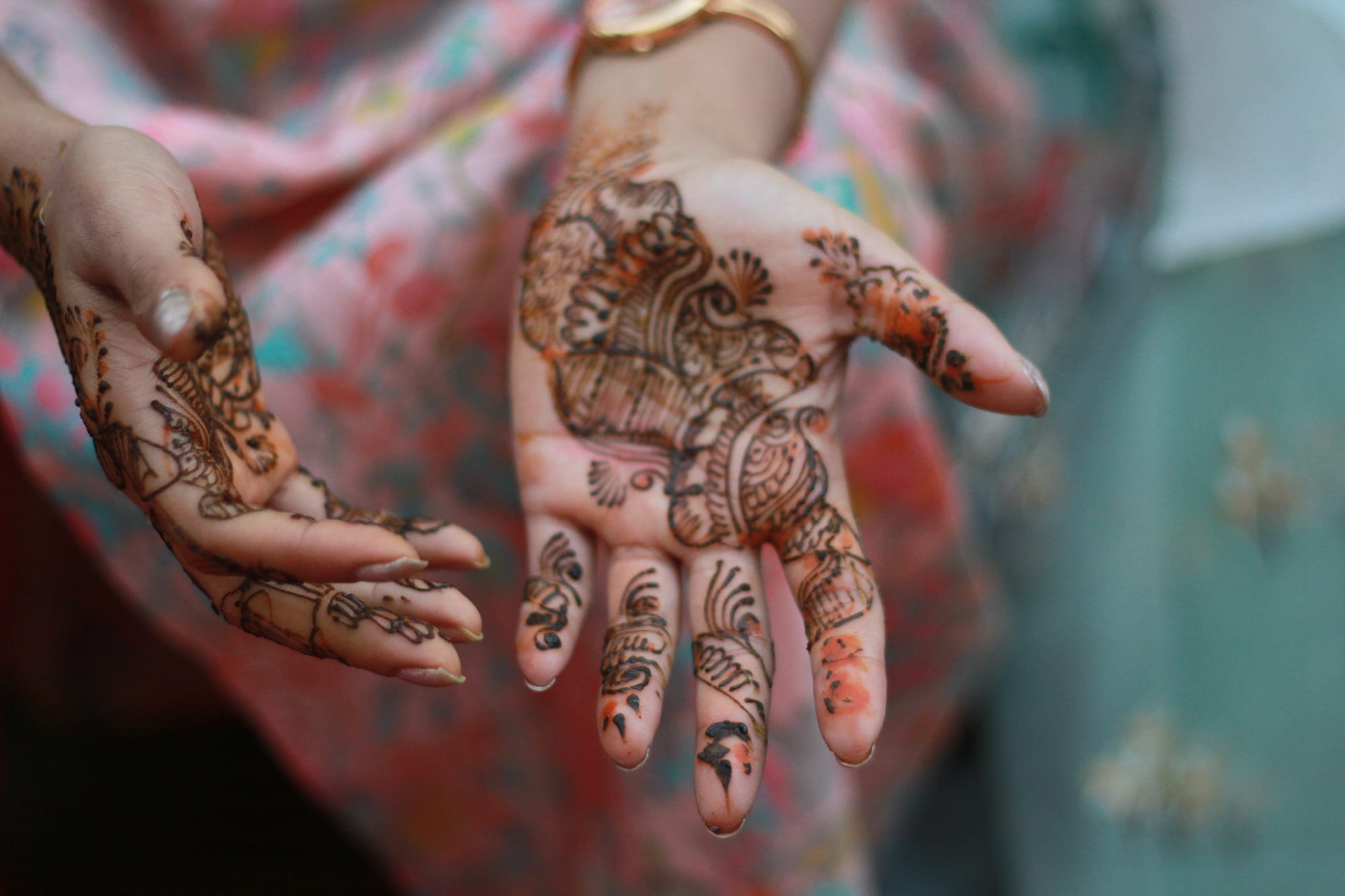 Two open hands covered in intricate dark brown henna patterns of flowers and paisleys, held out against a pink and patterned garment