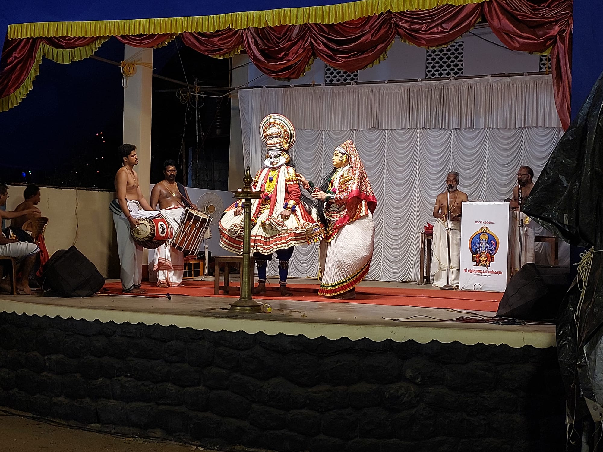 Two Kathakali performers in towering headdresses, painted faces and full white skirts stand on an outdoor stage at night beside a tall brass lamp, with drummers to one side and singers behind