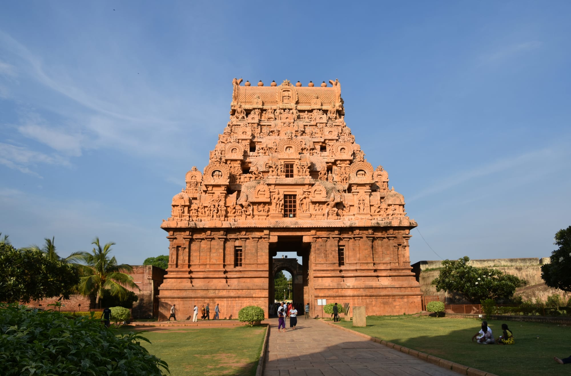 A tall, pyramid-shaped stone gateway tower covered in carved figures stands at the end of a paved path between lawns, with visitors walking through its entrance under a blue sky