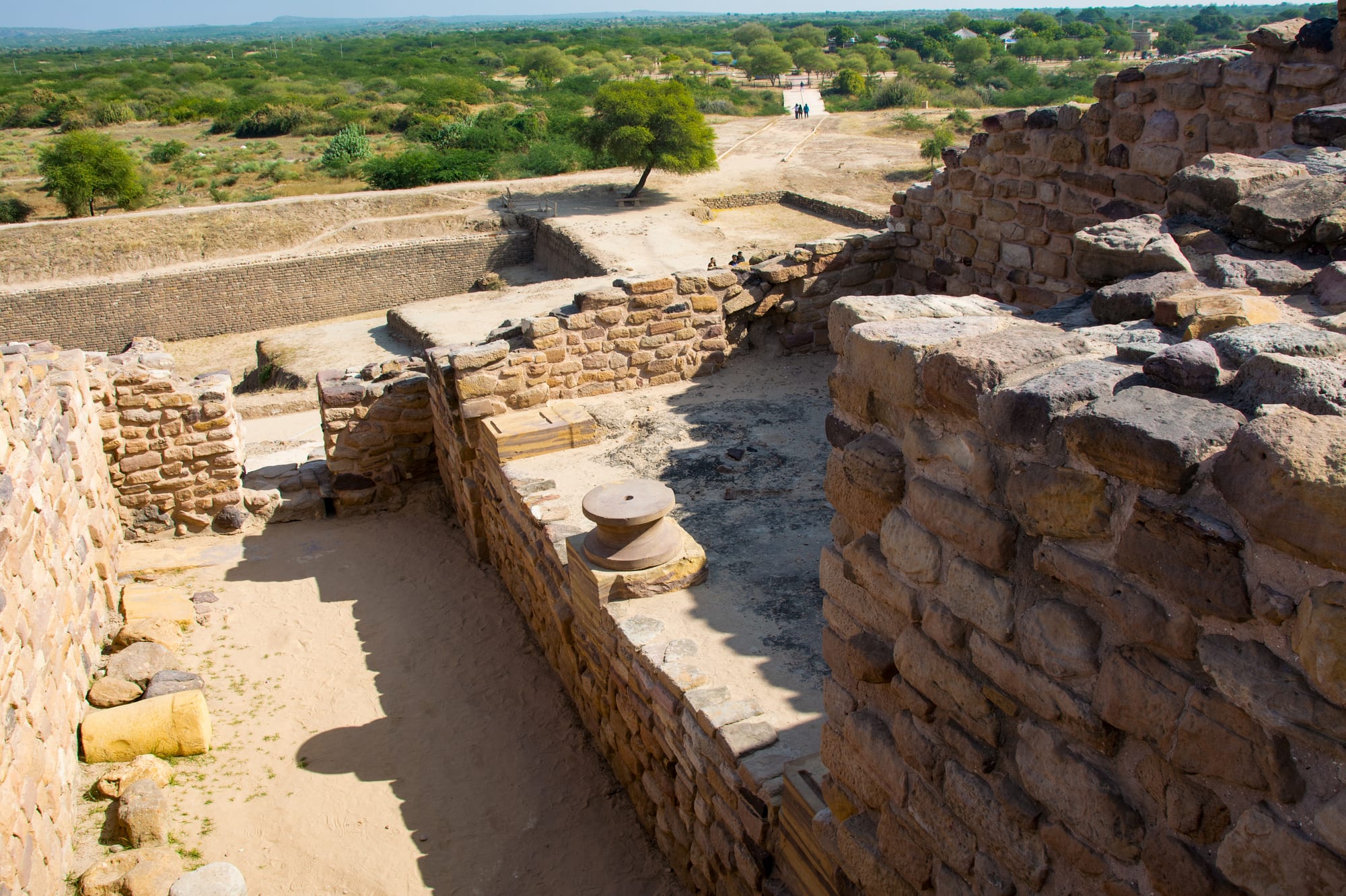 Excavated dry-stone walls, rooms and a round stone pillar base at an ancient site, with a large rectangular sunken enclosure and scrubland beyond