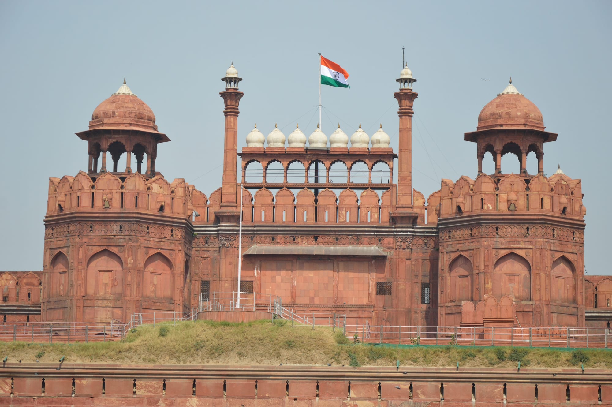 A massive red sandstone fortress gate with two domed octagonal towers, a row of small white domes and slender minarets, and the Indian tricolour flying from a flagpole above it