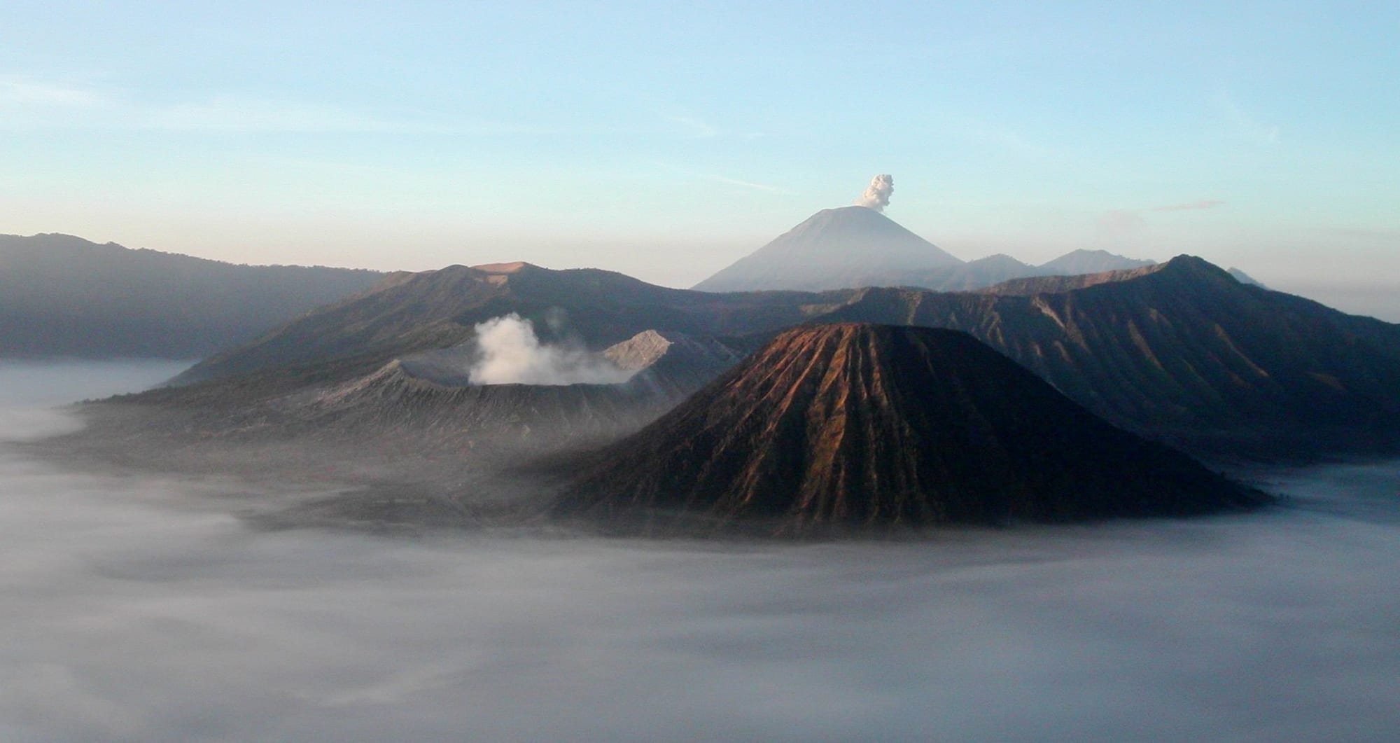 A smoking crater cone rising from a flat grey sea of sand, with a taller volcano behind at sunrise