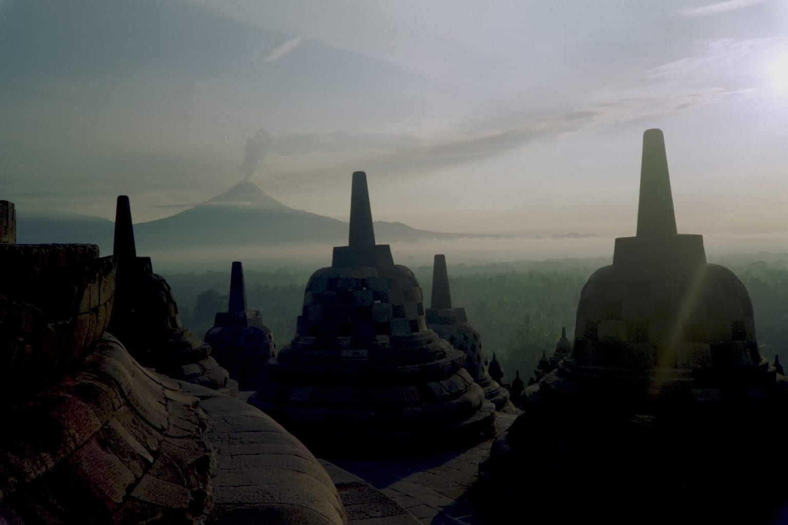 Rows of bell-shaped stone stupas on a temple terrace with a volcano in the mist beyond
