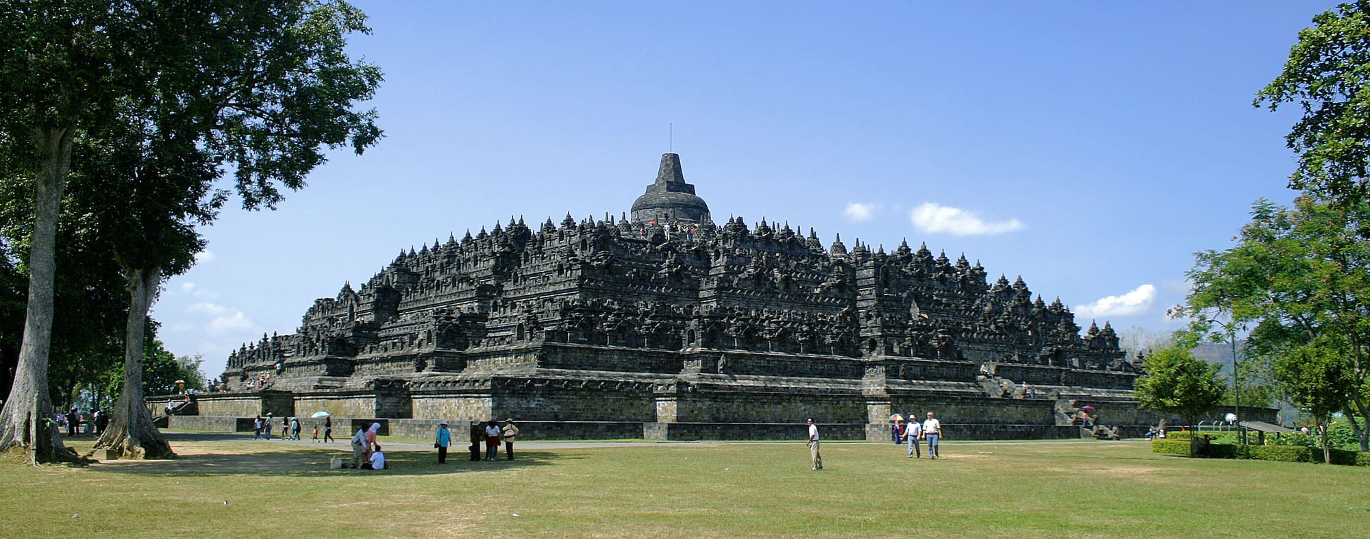A massive stepped stone pyramid temple with bell-shaped stupas, jungle and volcano behind
