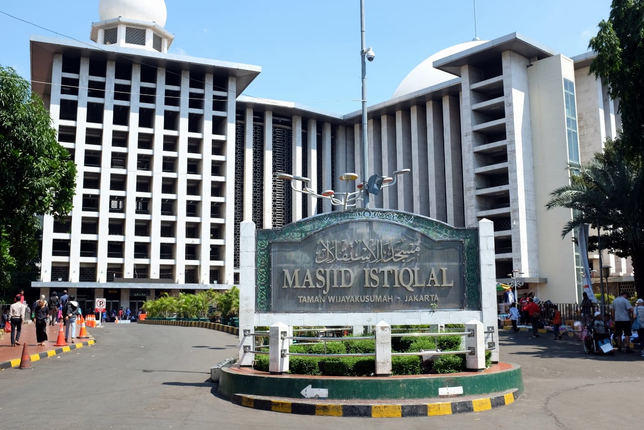A large white modernist mosque building with a dome behind it, fronted by a green and white sign reading Masjid Istiqlal in Arabic and Latin script