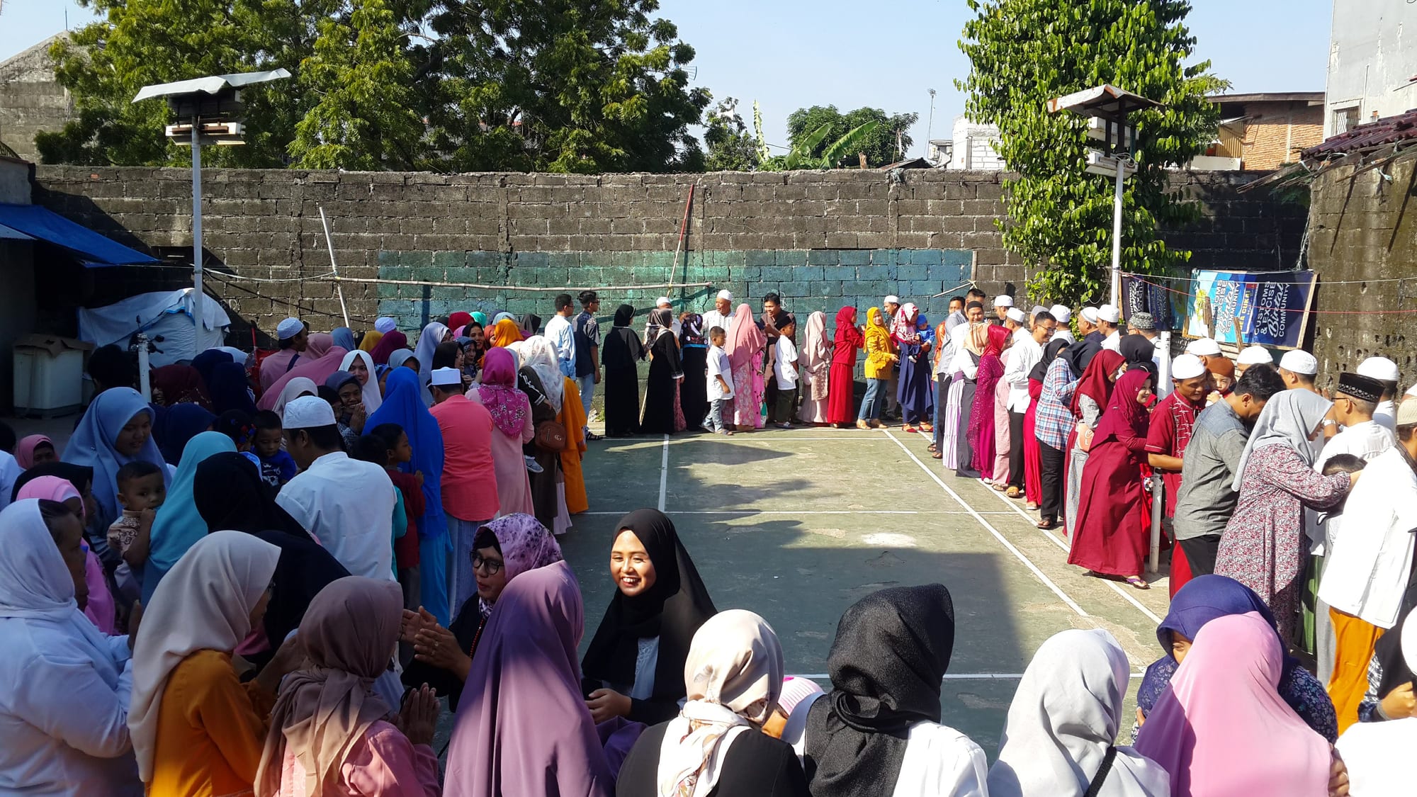 A long line of people, many women in headscarves and men in white caps, filing around an open-air court as they greet one another