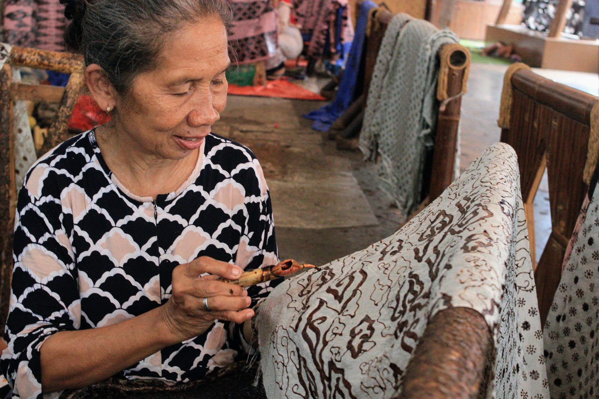 An older woman in a black and white patterned blouse holds a small copper-spouted tool, drawing wax lines onto cloth draped over a wooden frame