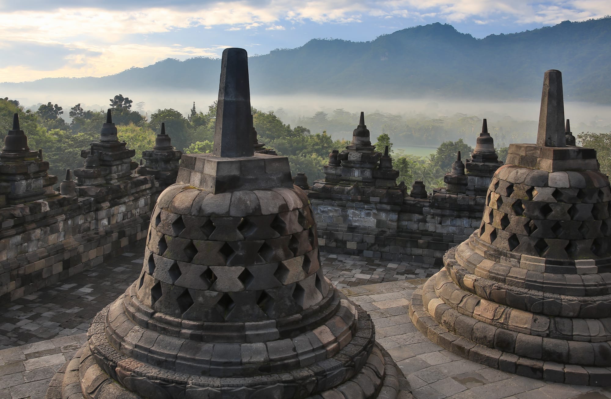 Rows of bell-shaped stone stupas on the upper terraces of a vast stepped temple, volcanoes behind