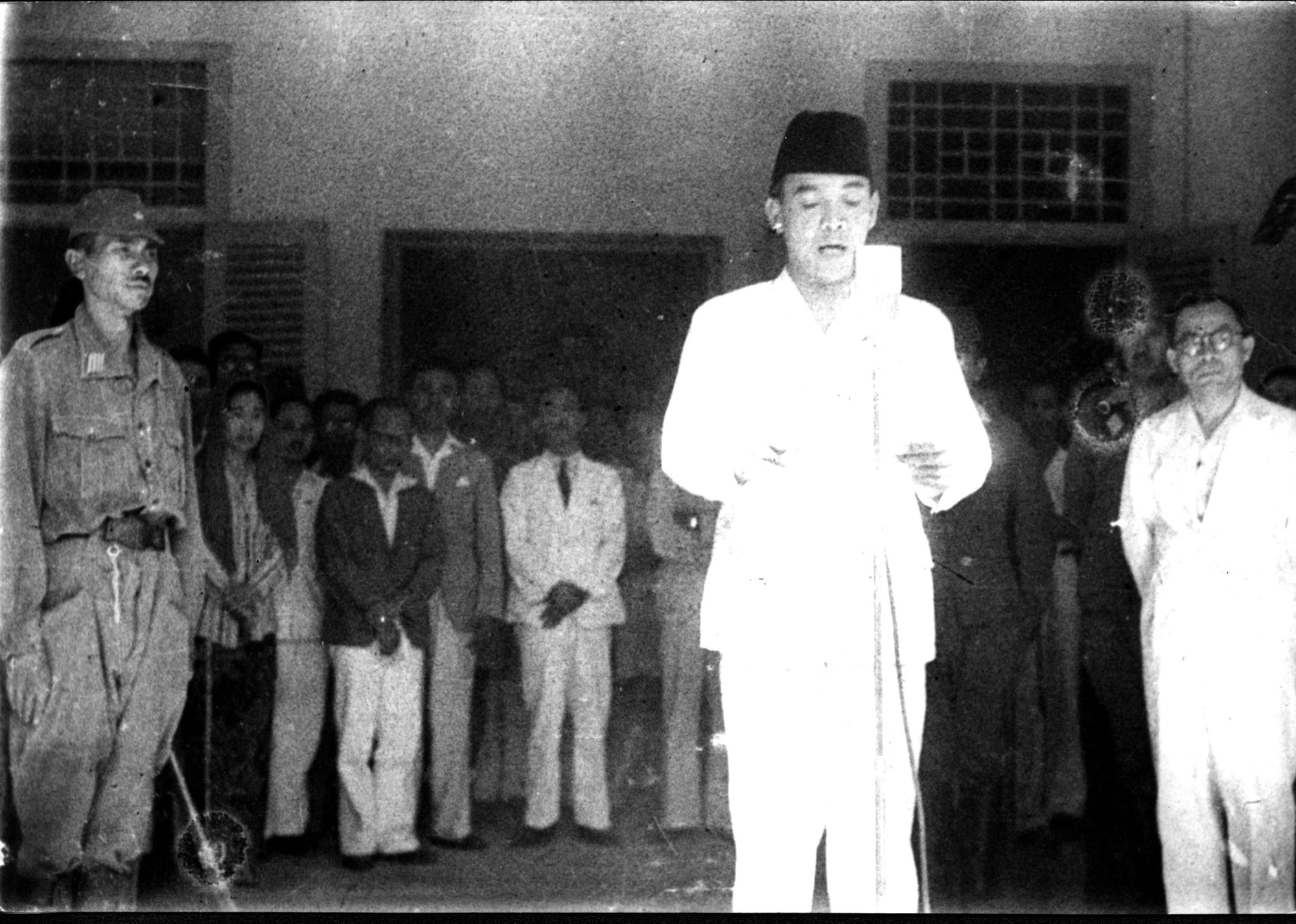 Sukarno reading the proclamation of independence from a document before a small crowd, 1945