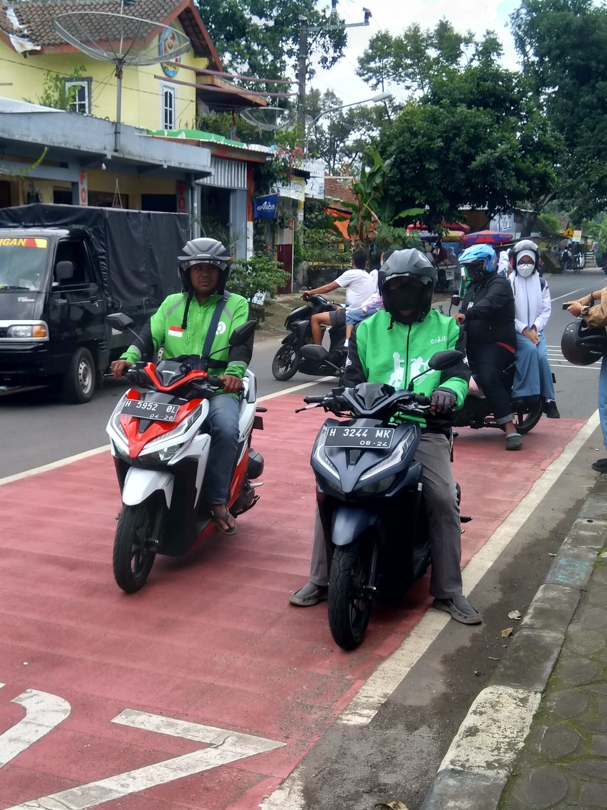 Two helmeted Gojek motorbike-taxi riders in green jackets waiting on their scooters at a roadside stand