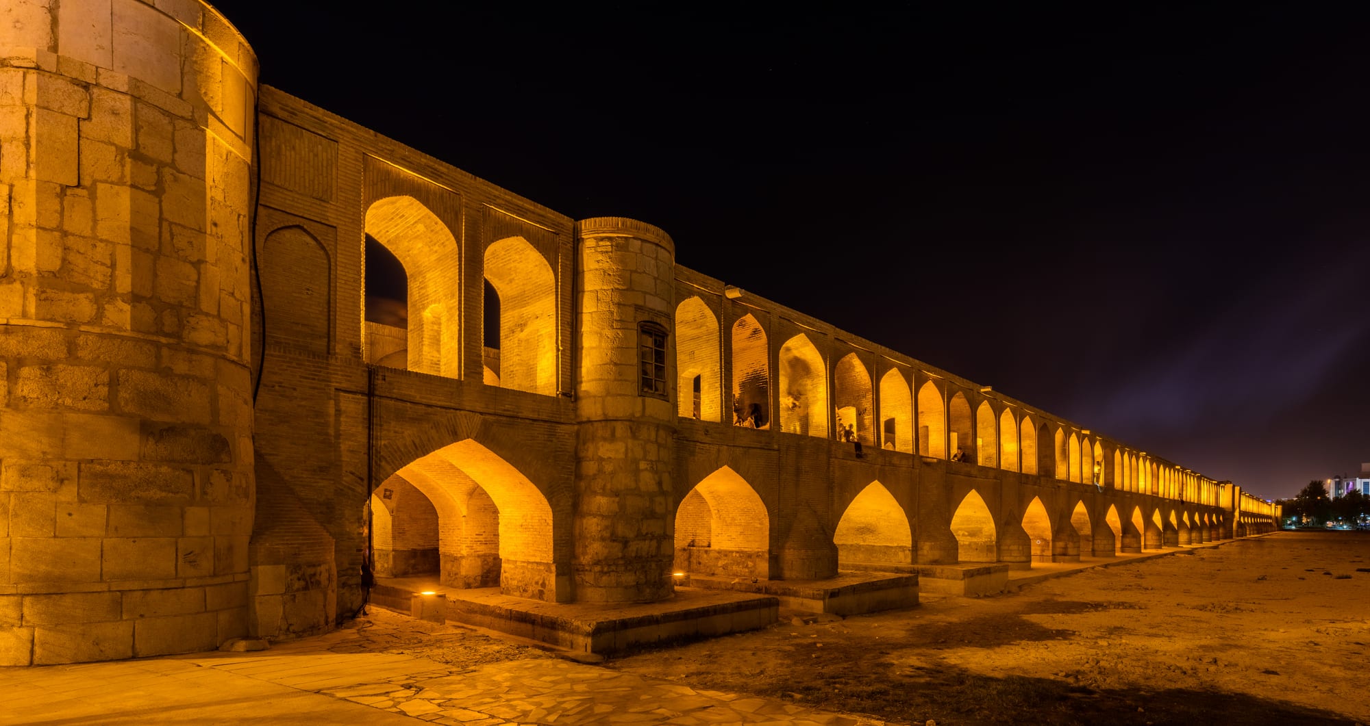 A long two-tier arched stone bridge spanning a wide riverbed at dusk, lit up