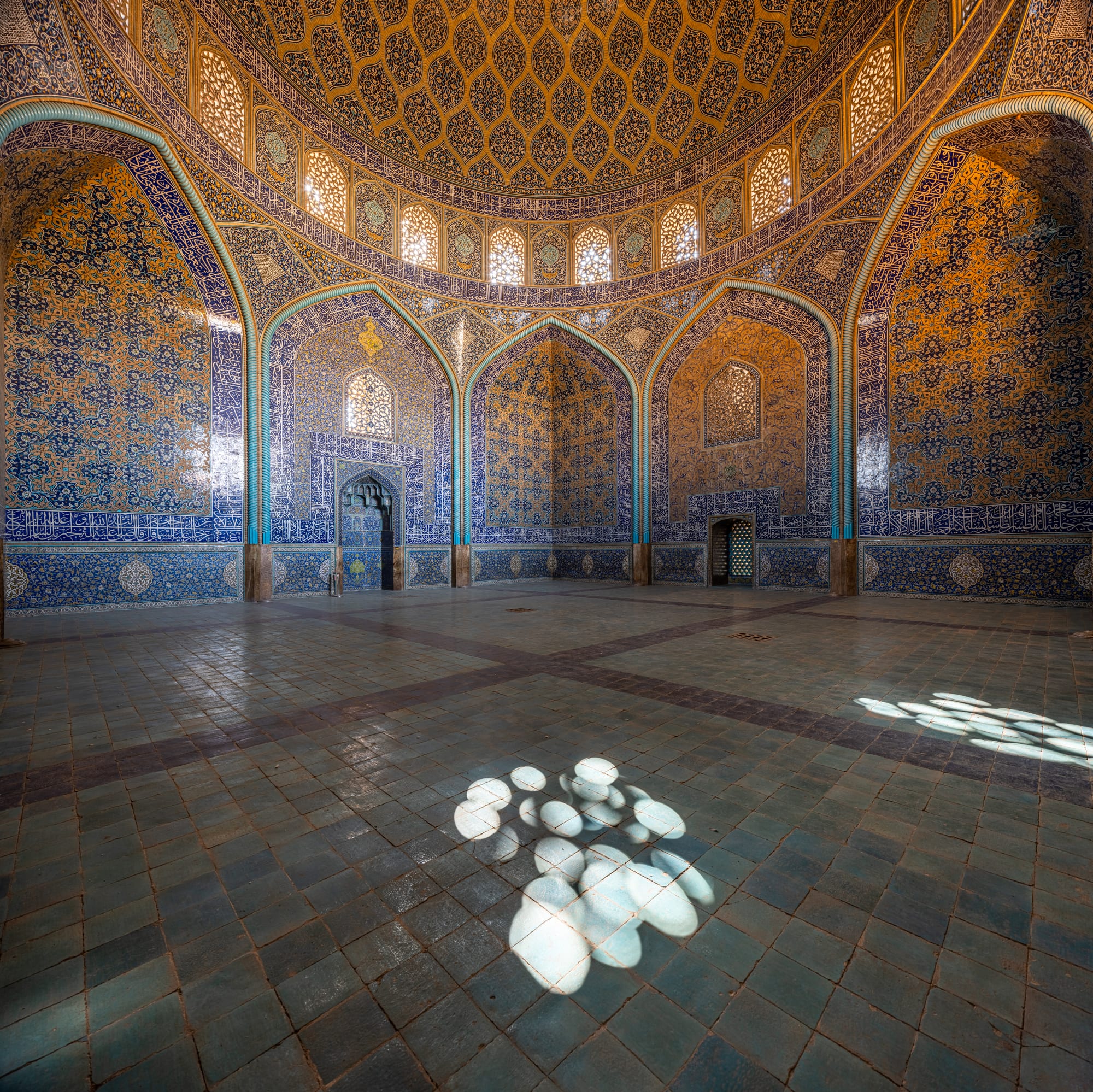 A domed mosque interior of intricate blue, cream, and gold tilework rising to a central medallion