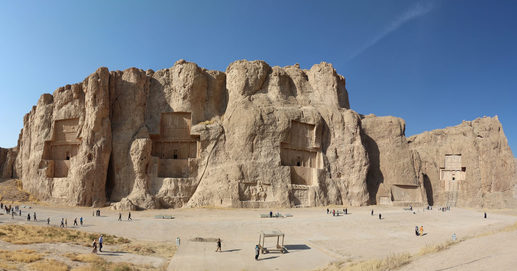 A long sandstone cliff with four cross-shaped tomb facades cut high into the rock, and visitors walking on the open ground below