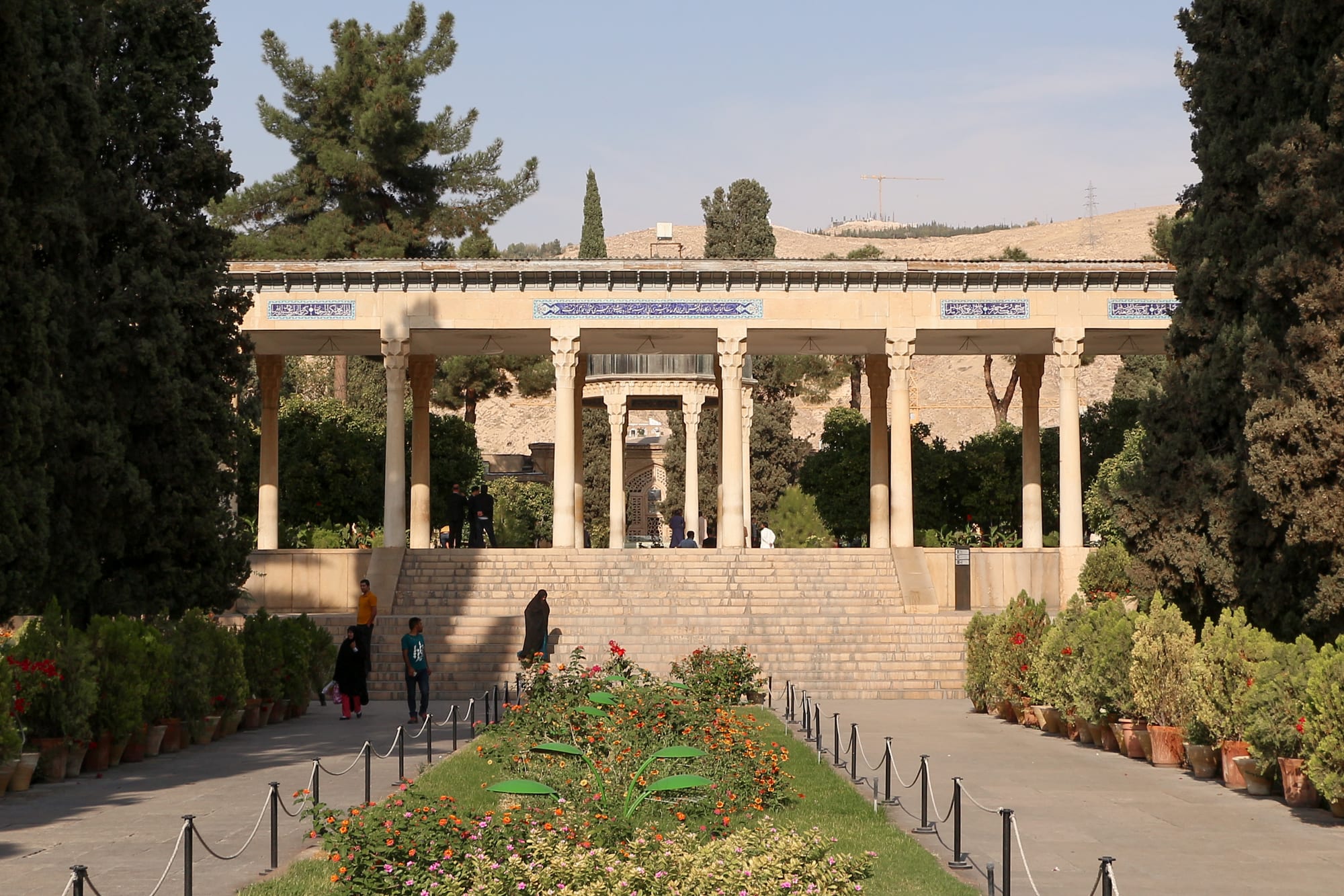 A pillared stone pavilion at the top of a broad flight of steps, approached by a paved path lined with potted plants and flowerbeds, with tall cypresses on either side