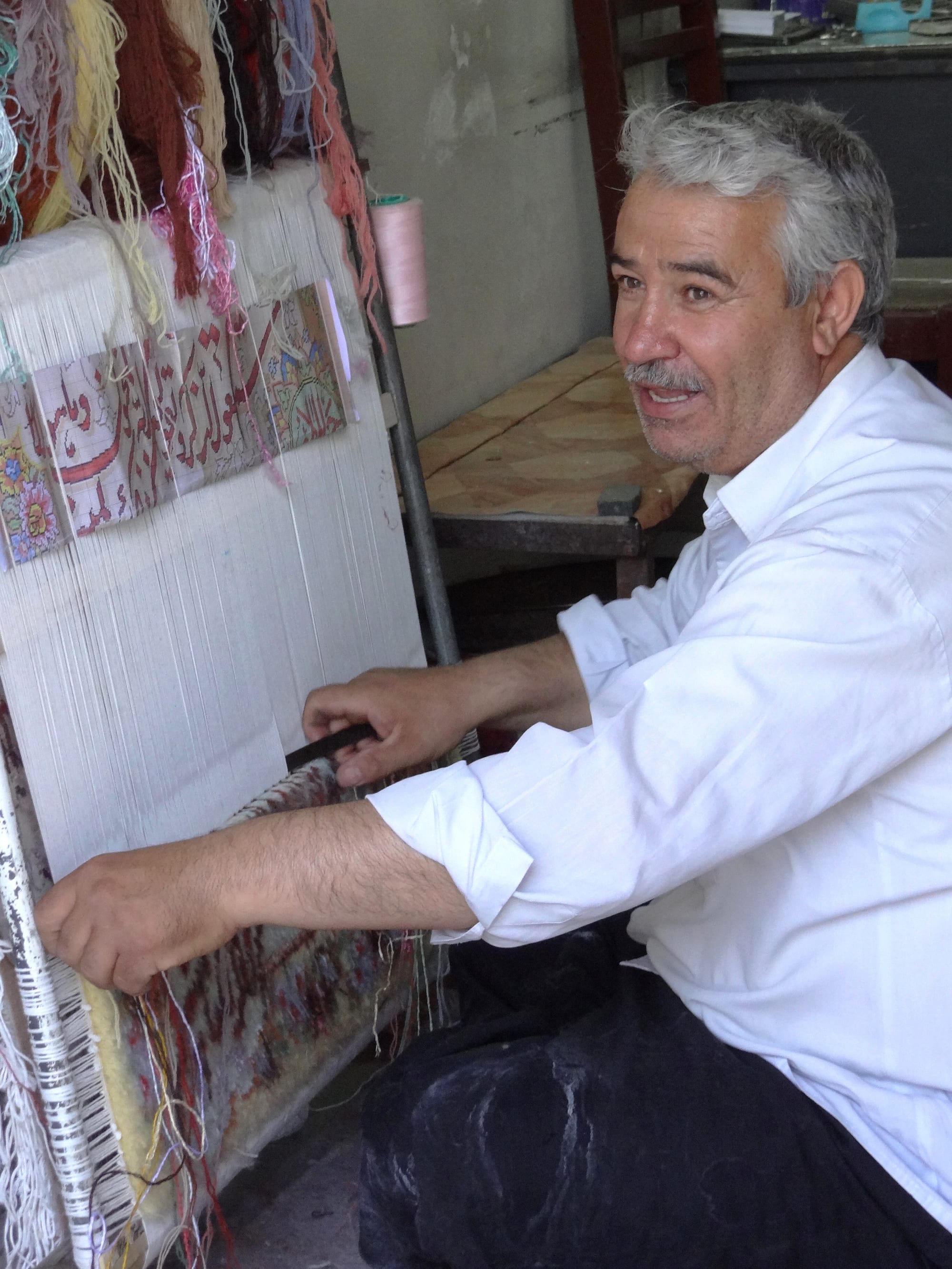A grey-haired man in a white shirt sits at an upright loom, pulling a tool through the warp threads, with coloured yarns hanging above and a paper pattern pinned to the loom