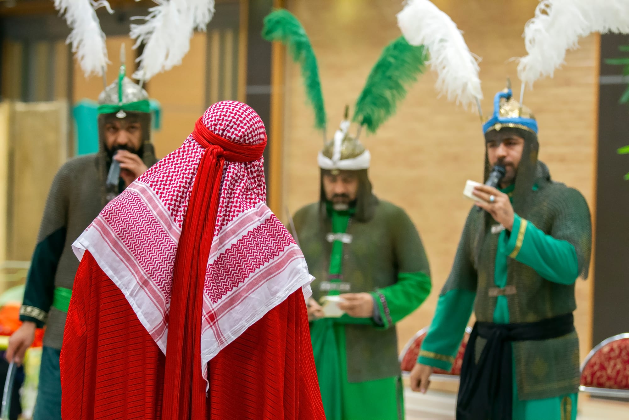 Performers in costume on an indoor stage, three men in chain mail and plumed helmets singing into microphones, with a figure in a red robe and red and white headscarf in the foreground