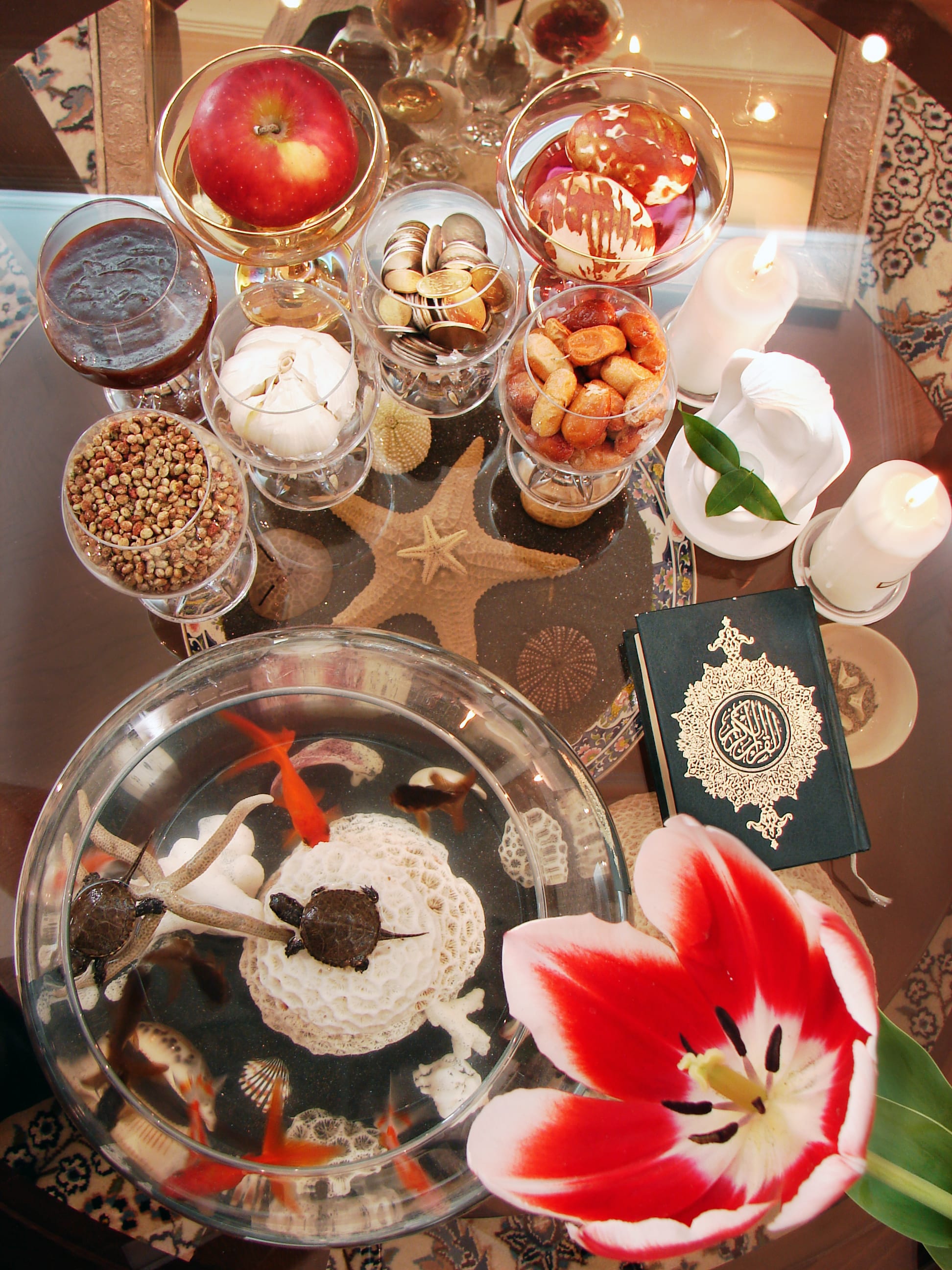 A Nowruz haft-sin table seen from above, with an apple, garlic, coins, painted eggs, a bowl of goldfish, candles, a copy of the Quran and a red and white tulip