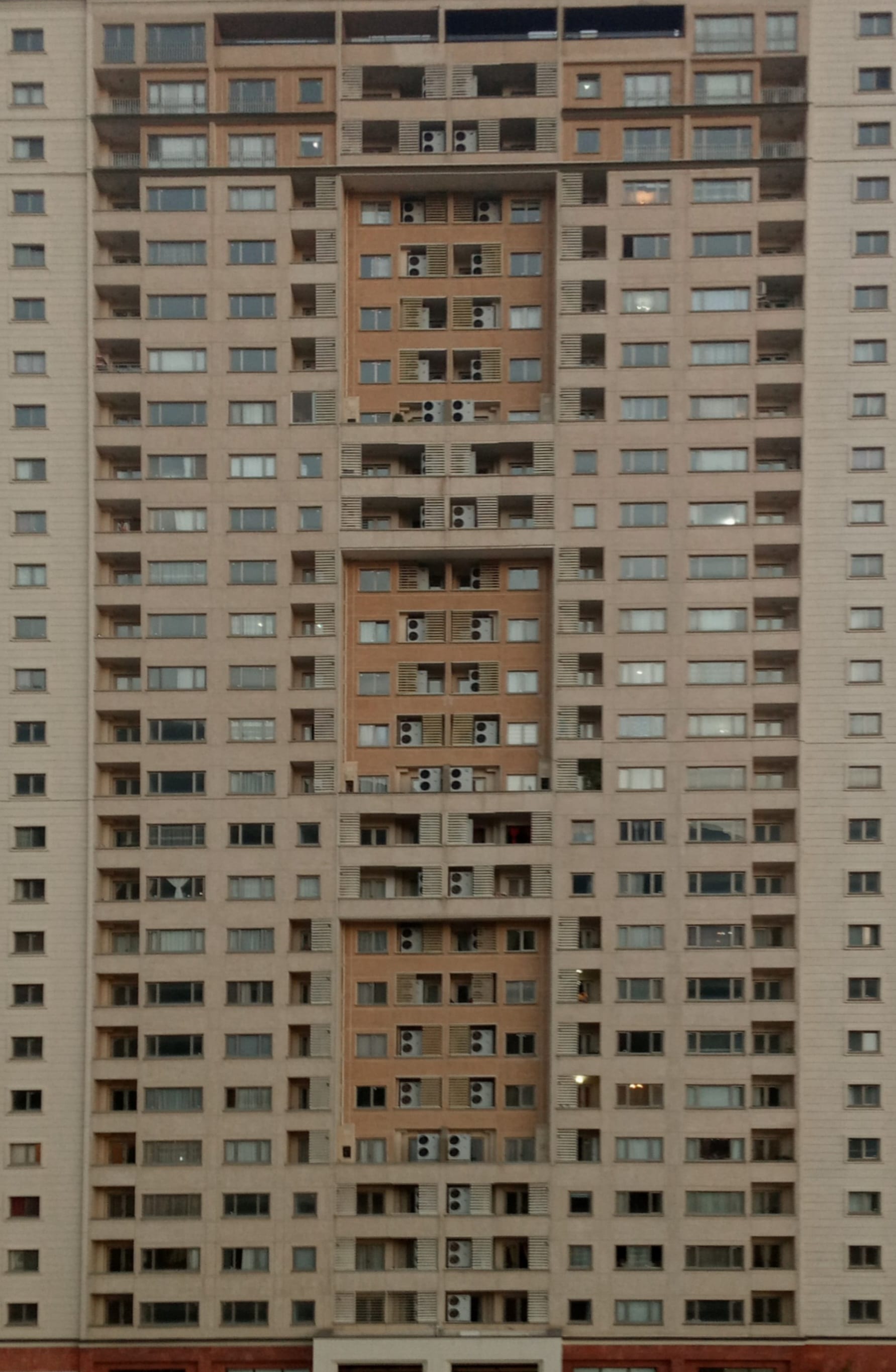 A tall Tehran residential tower at dusk, its facade a dense grid of apartment windows, balconies and air-conditioning units