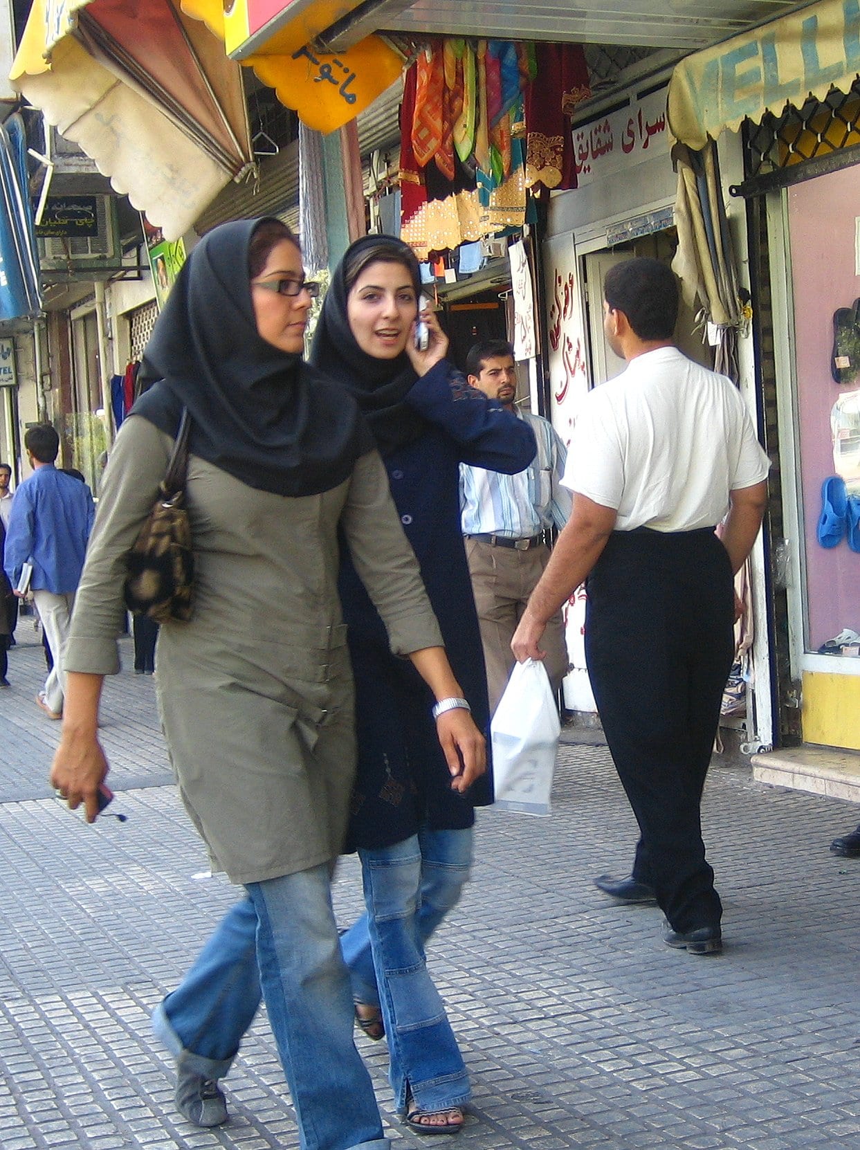 Two young women in headscarves and jeans walking along a shop-lined street, one talking on a mobile phone