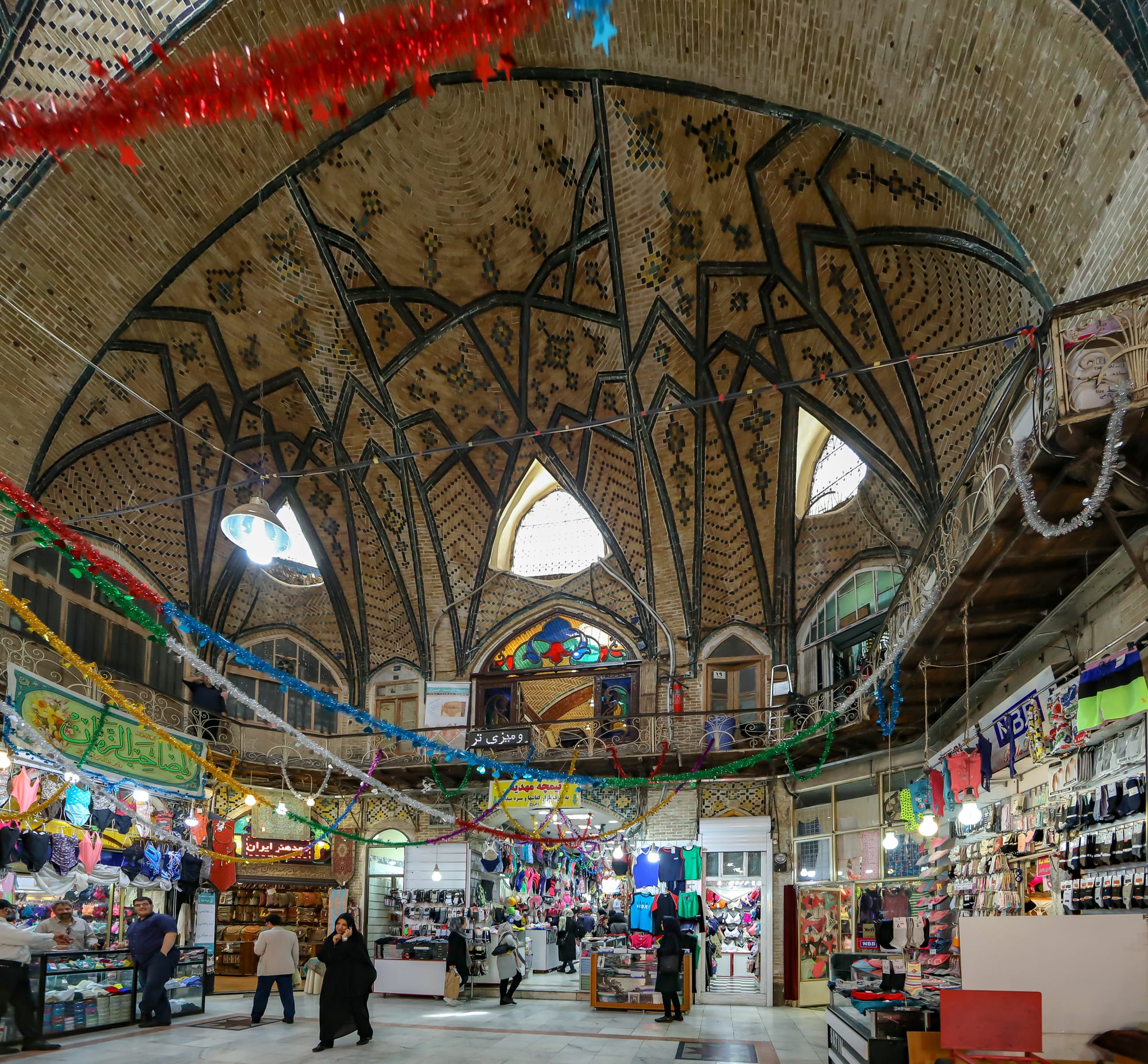 Shoppers and vendors beneath the vaulted brick ceiling of Tehran's Grand Bazaar, its arches hung with coloured streamers