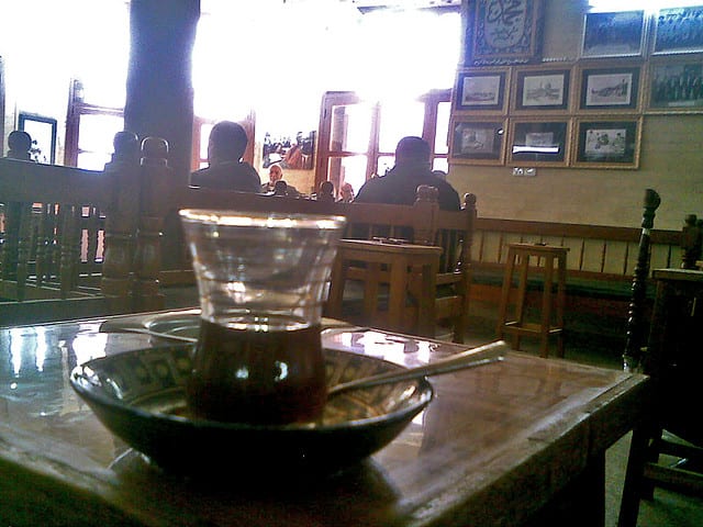 A glass of tea on a saucer on a wooden table inside a dim, historic Baghdad coffeehouse, with patrons seated in the background