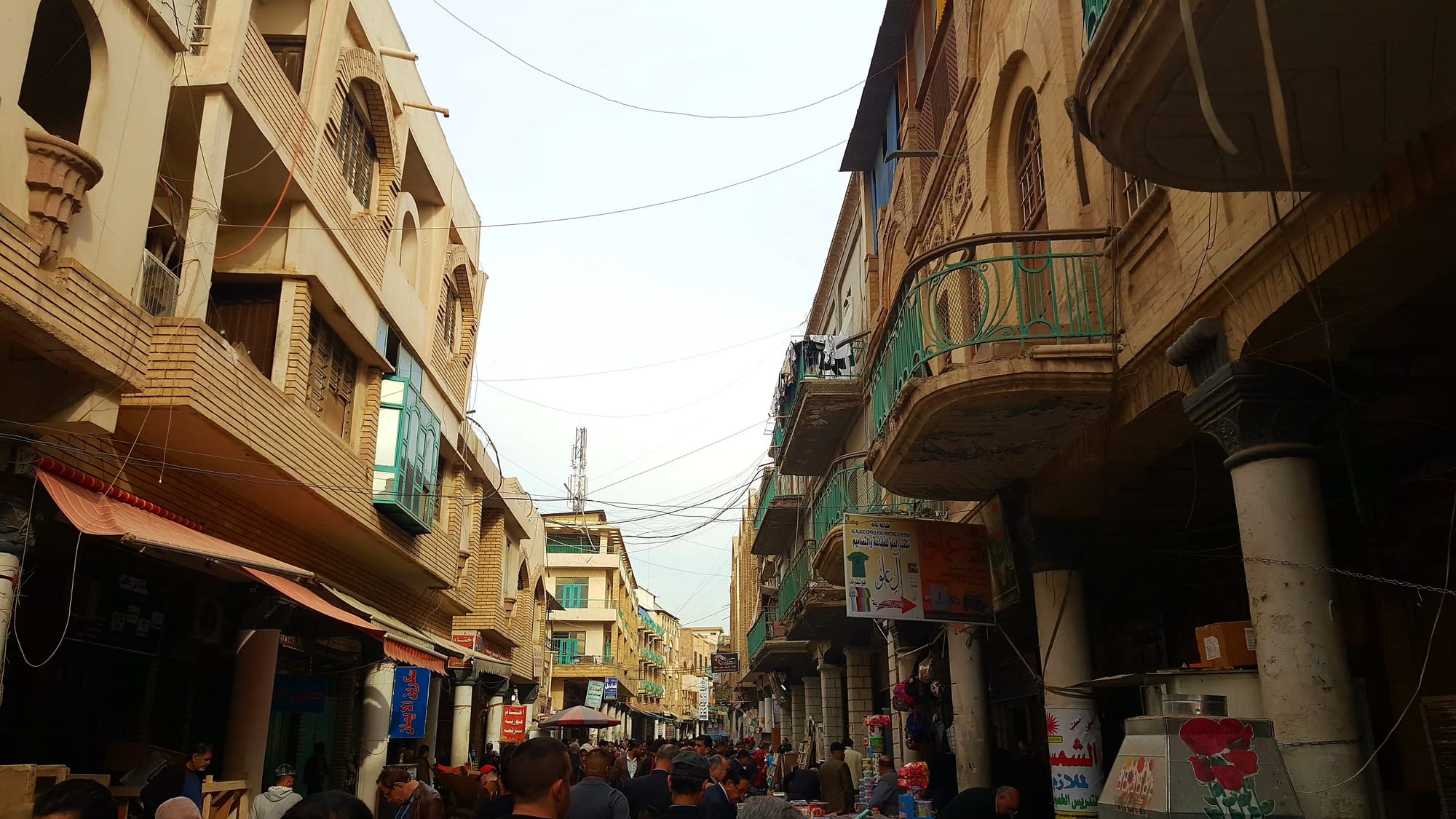 A crowded street in Baghdad's book-market district, tall balconied buildings on both sides strung with overhead cables