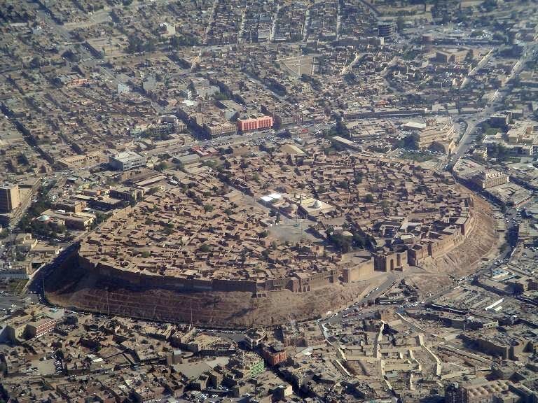 A walled ancient citadel of packed earth buildings rising on a mound above a modern city