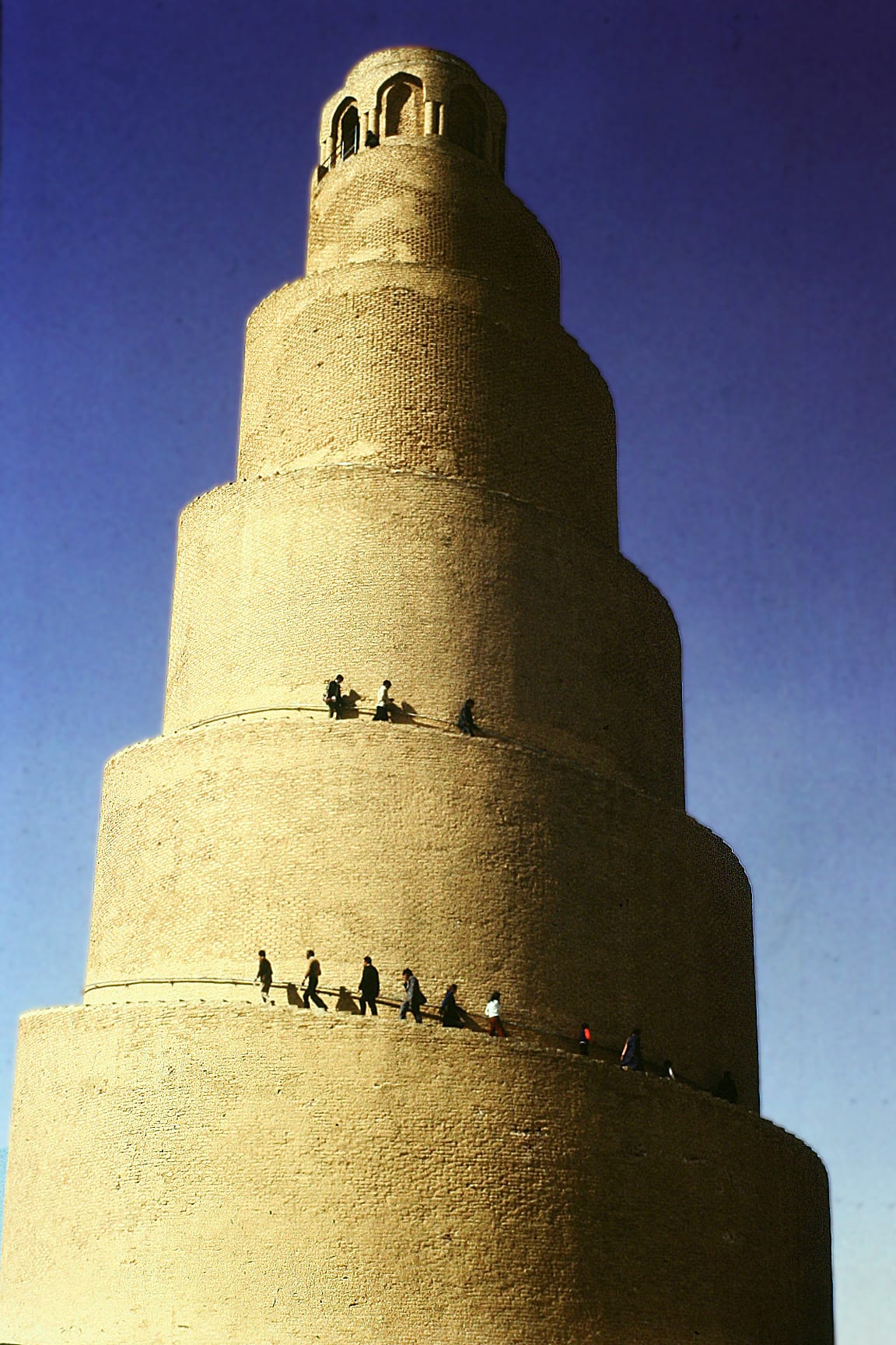 A close-up view of a spiralling brick minaret with a ramp winding up its cone shape, tiny figures climbing it