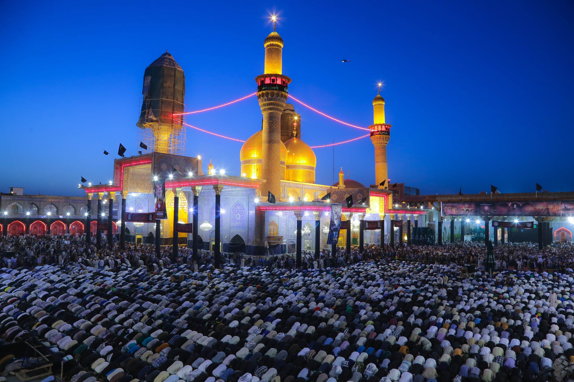 Golden domes and two tall golden minarets rising above an illuminated colonnade at dusk, with a vast crowd of worshippers kneeling in rows across the courtyard in front