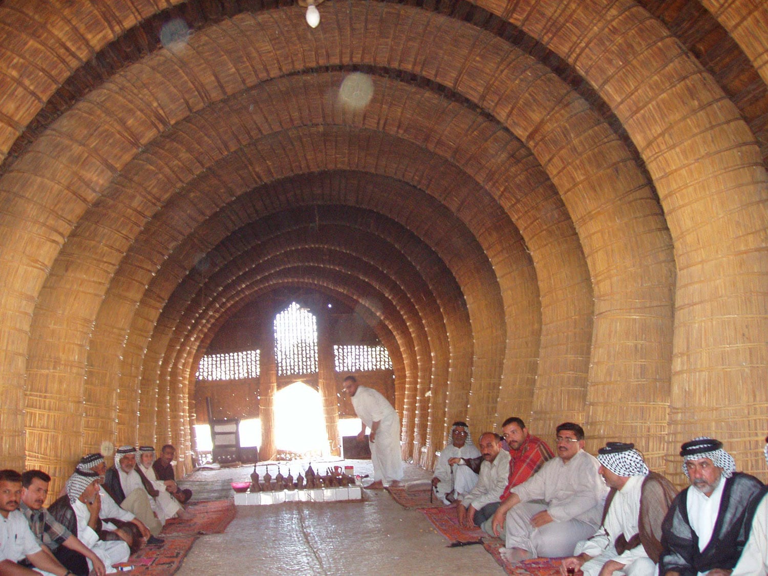 Men in white robes and chequered headscarves seated on rugs along both sides of a long hall built of arched reed bundles, with a row of coffee pots set out in the middle of the floor