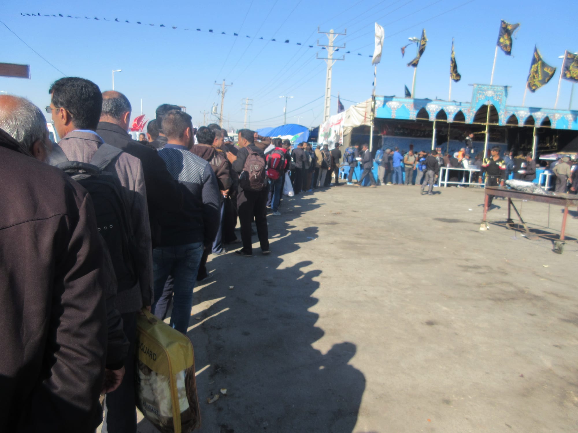 A long queue of men in jackets waiting along a dusty roadside towards a blue decorated stall hung with black and gold banners, under strings of small flags