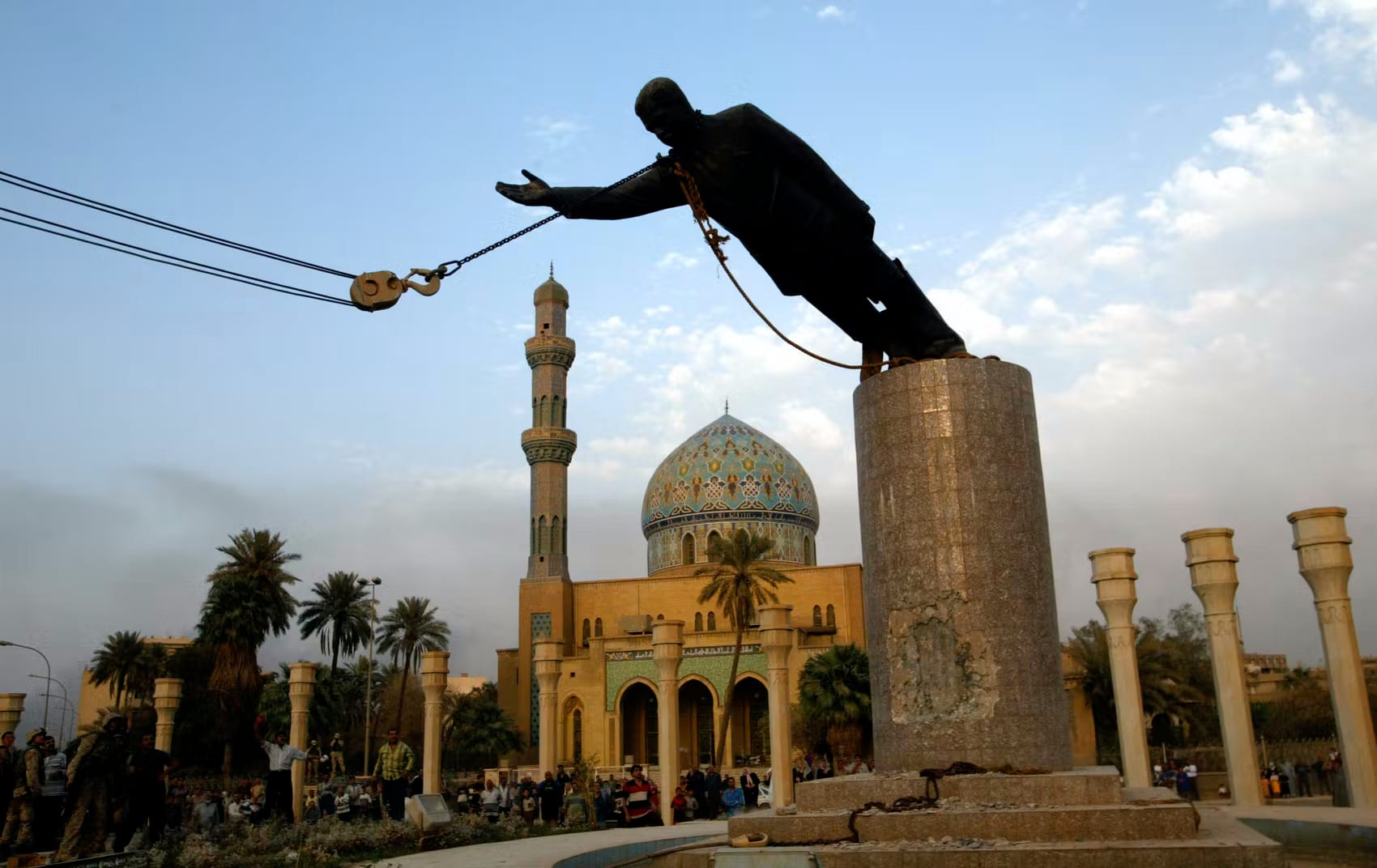 A large statue of Saddam Hussein being pulled down by a crowd and a US military vehicle in a Baghdad square