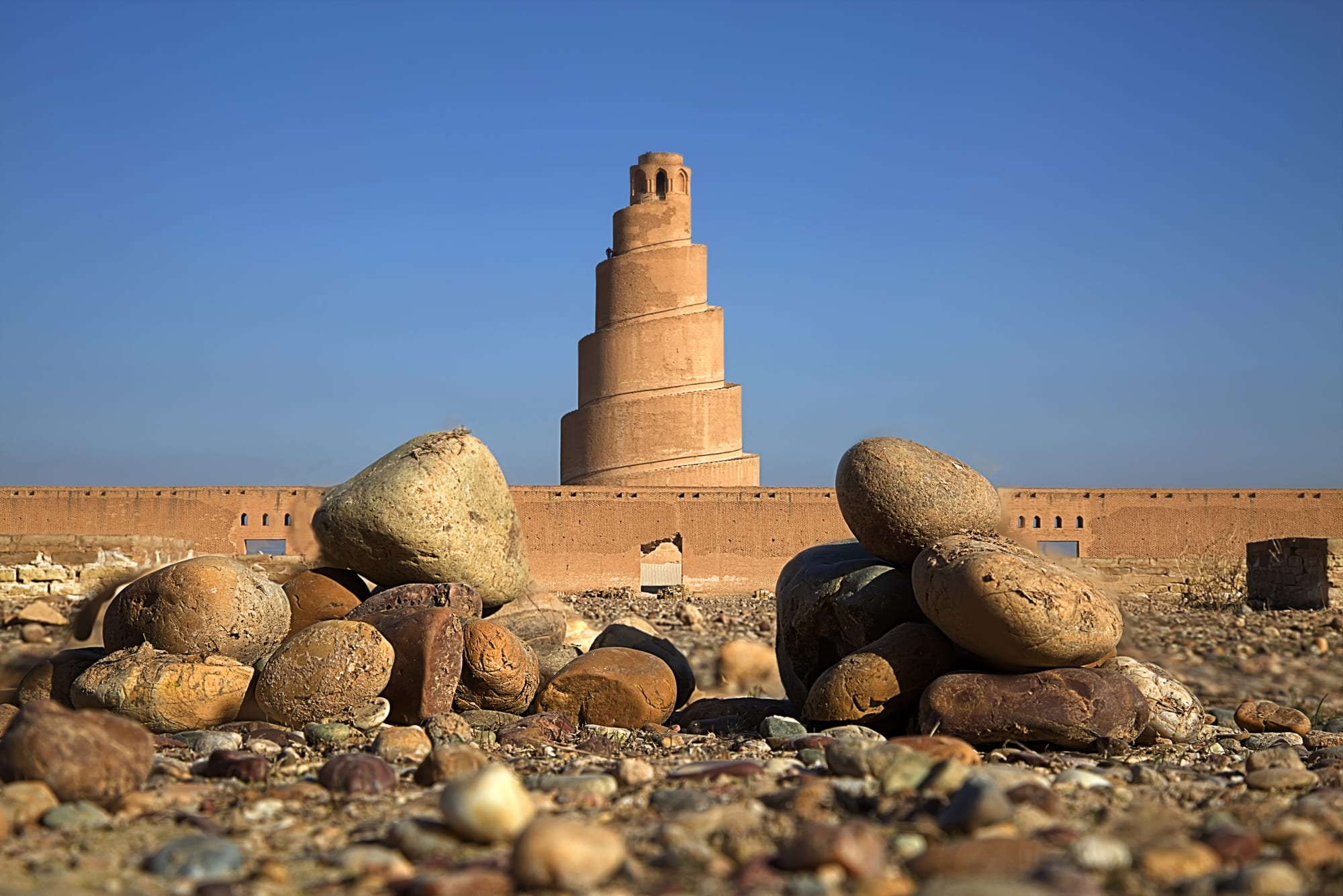 A tall spiral brick minaret rising behind a long buttressed mud-brick wall, with rounded river stones in the foreground under a clear blue sky