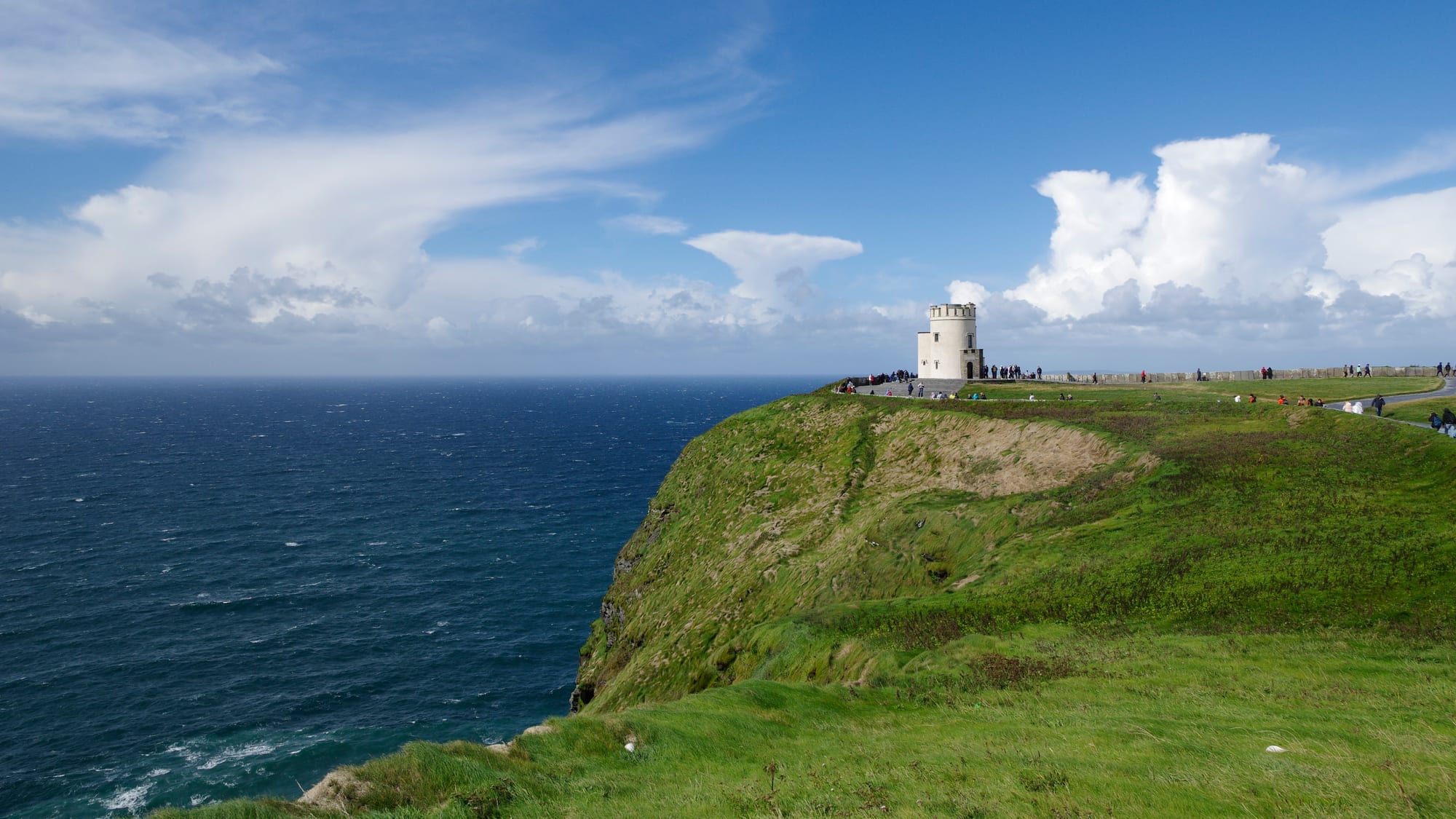 A person on a clifftop path with immense layered sea cliffs stretching into the distance