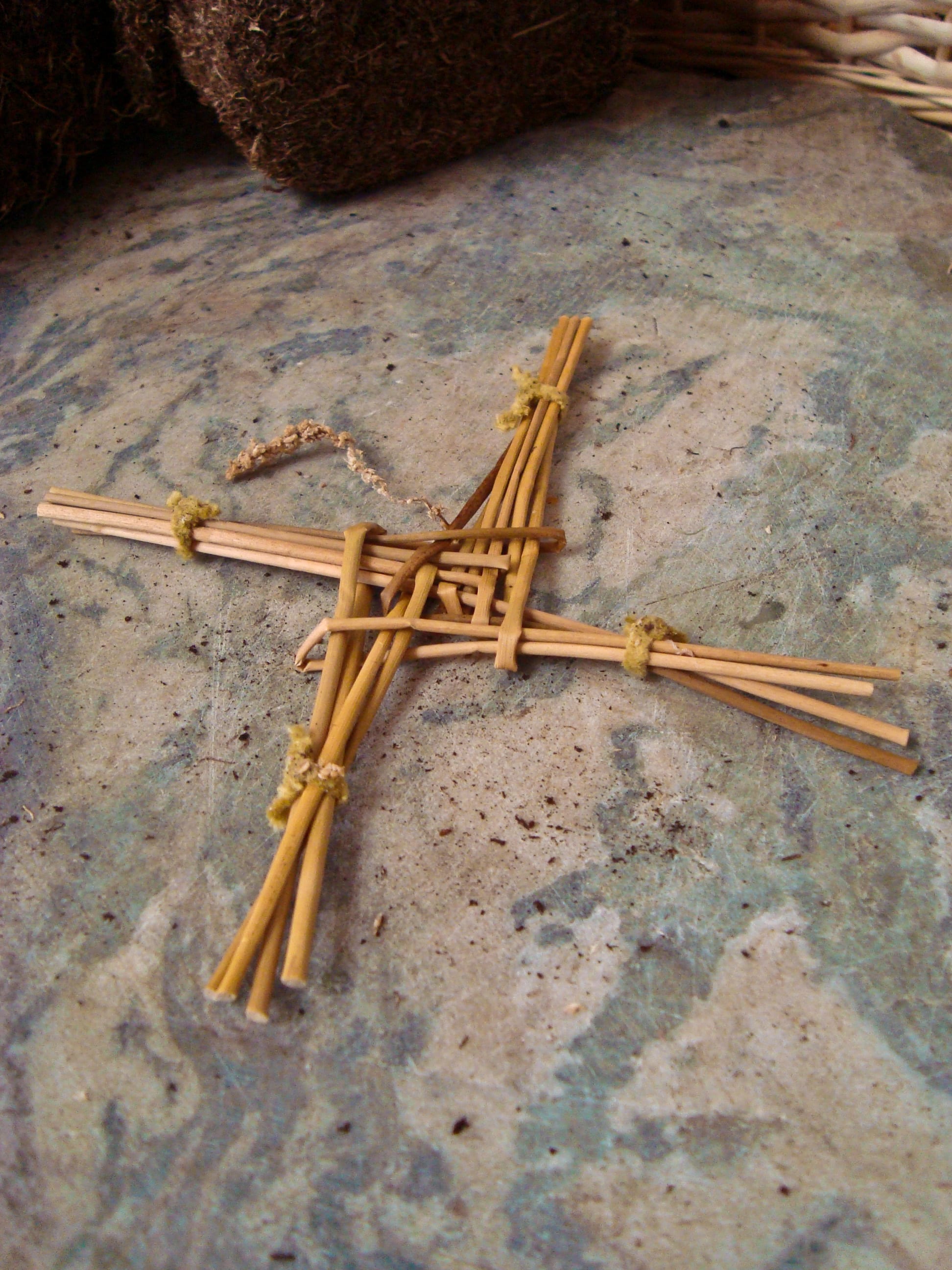 A four-armed cross woven from pale straw-coloured stems, the ends of each arm tied, resting on a flat stone surface
