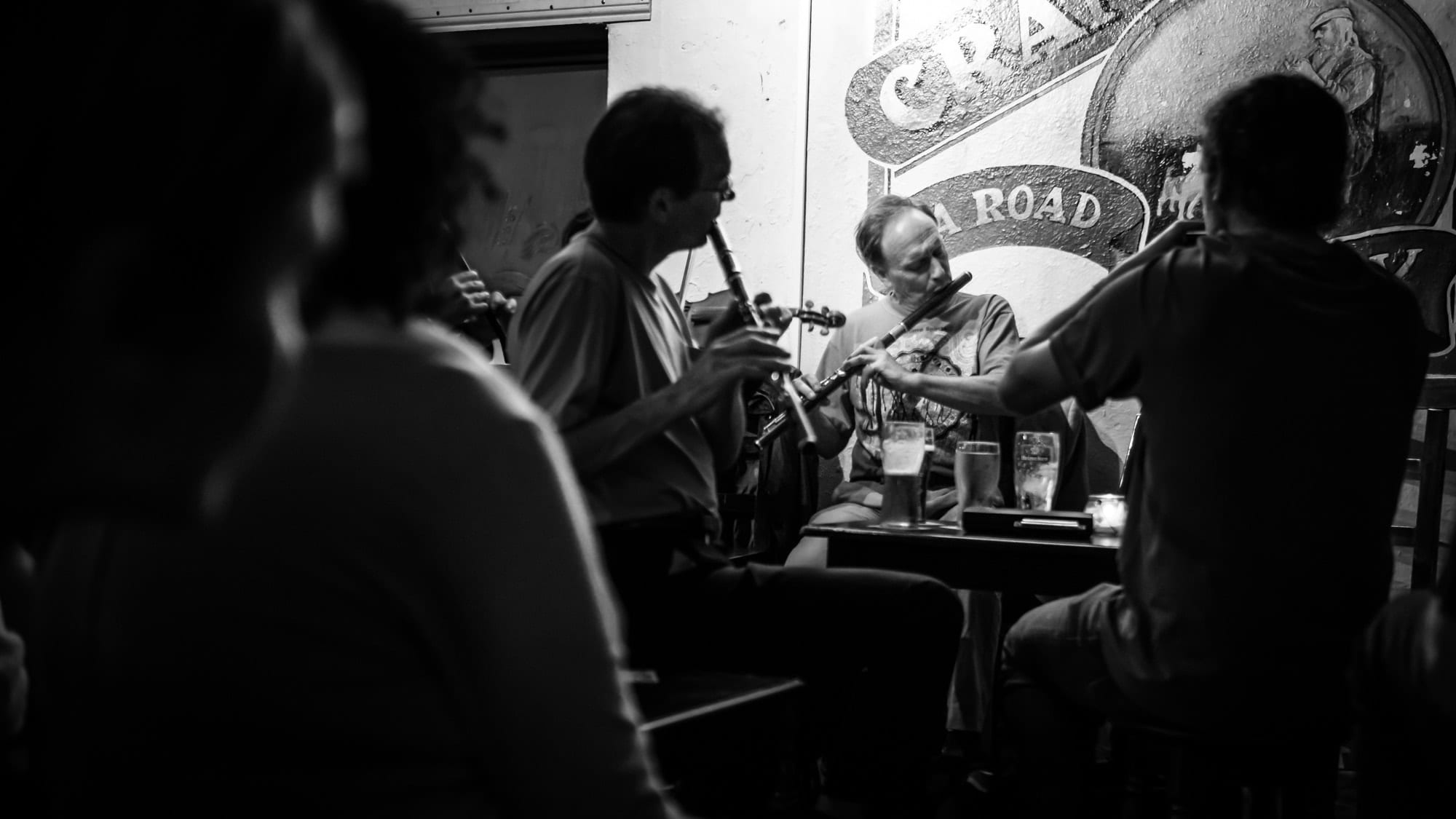 Black and white photograph of musicians seated around a small pub table playing flutes and a fiddle, with pints of stout on the table and listeners in the foreground