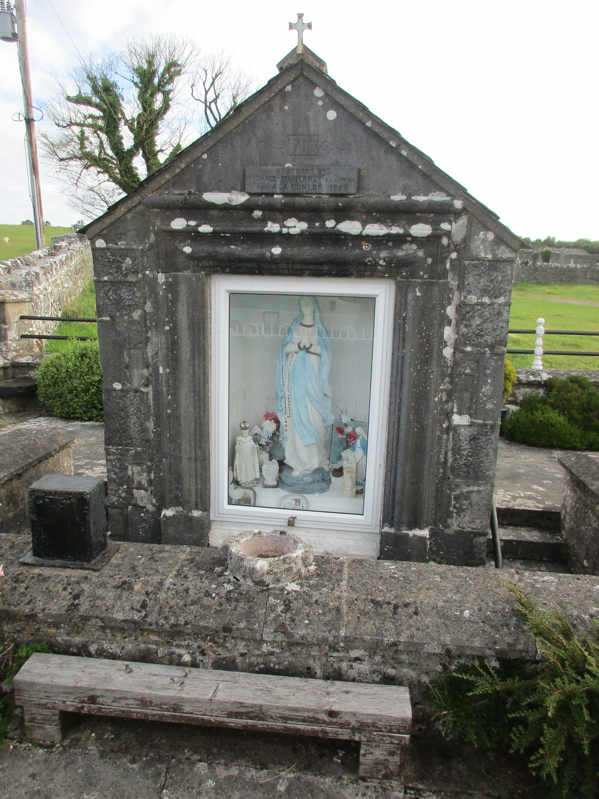 A small gabled stone well house topped with a cross, with a statue of the Virgin Mary and other figures displayed behind glass and a stone basin in front, set among fields and stone walls