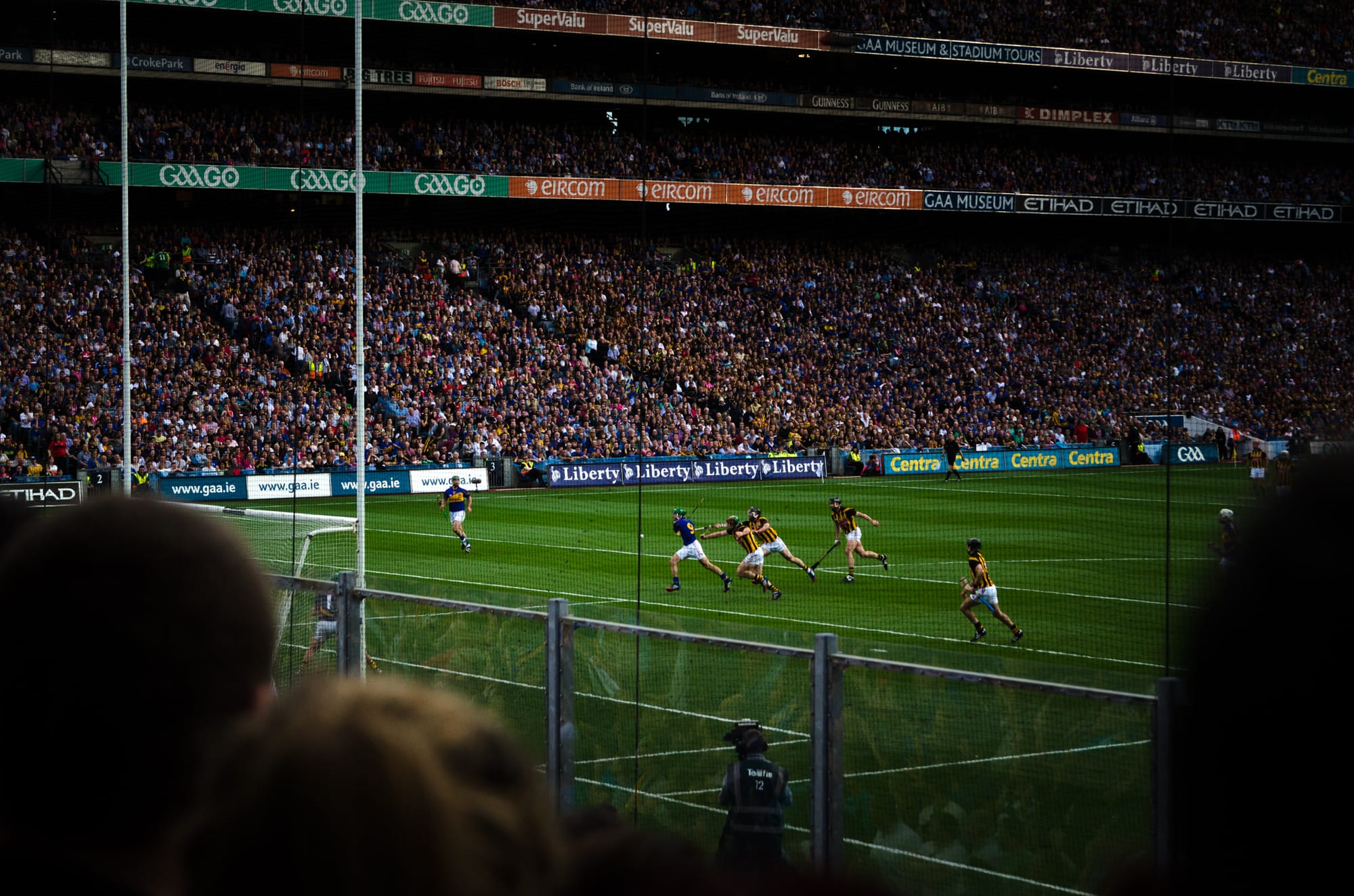 Hurlers in blue and in black and amber chasing the ball across the pitch in front of packed stands, seen from behind a fence at the edge of the field
