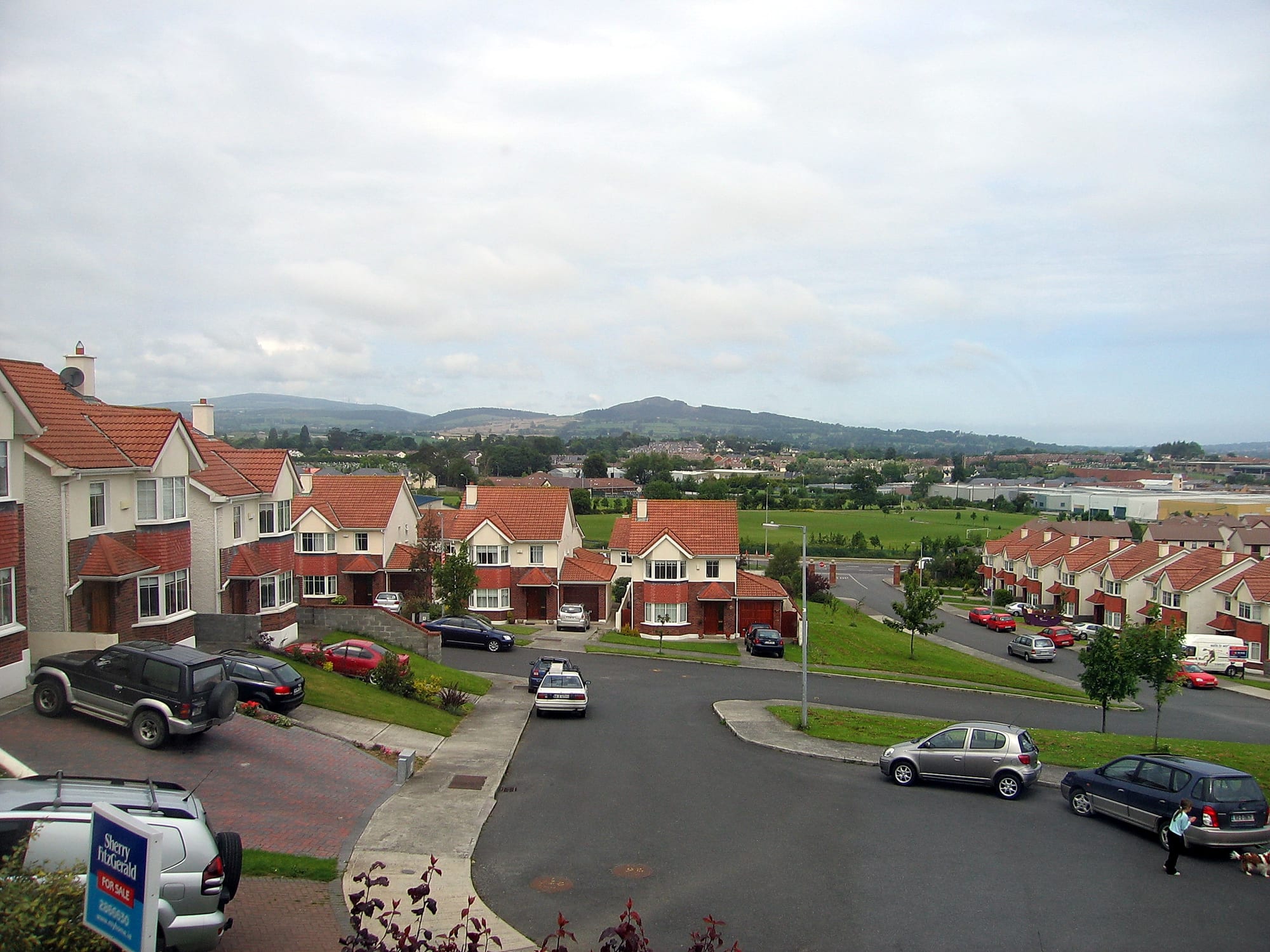A suburban Irish housing estate of red-roofed semi-detached houses seen from above, with parked cars and green hills in the distance