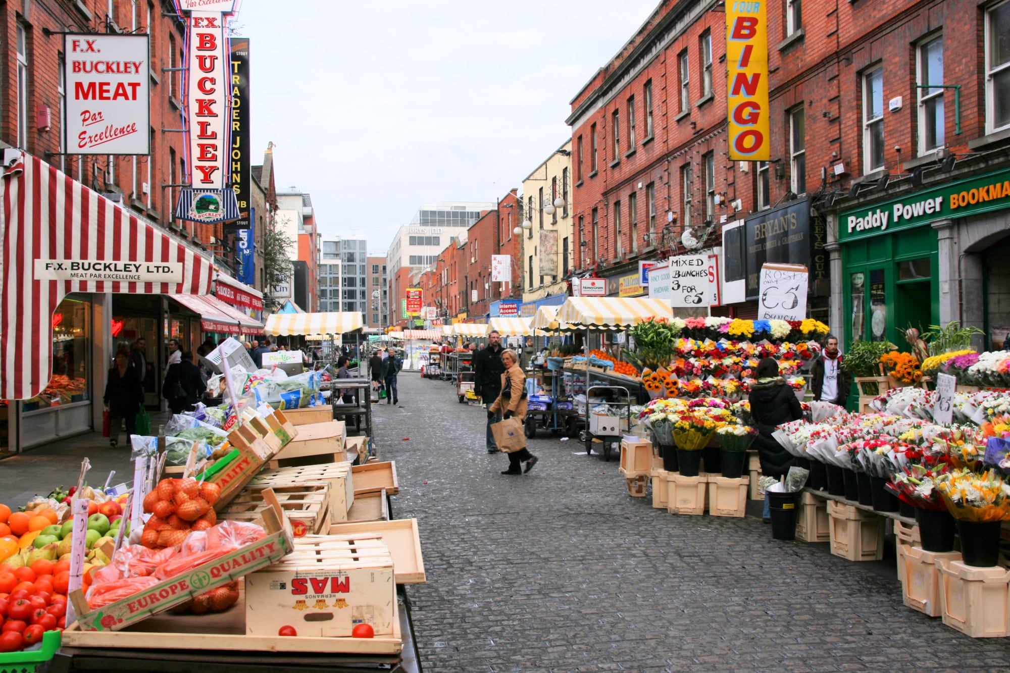 A cobbled Dublin street lined with fruit, vegetable and flower stalls under striped awnings, shoppers walking between them