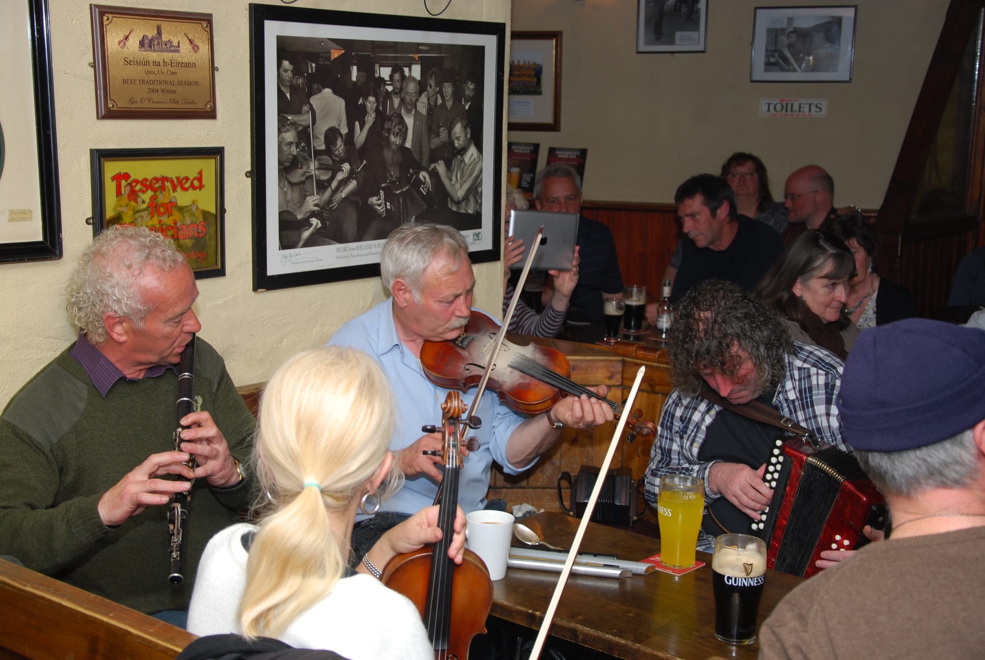 A group of musicians playing fiddle, clarinet and accordion around a table in a crowded, memorabilia-lined pub