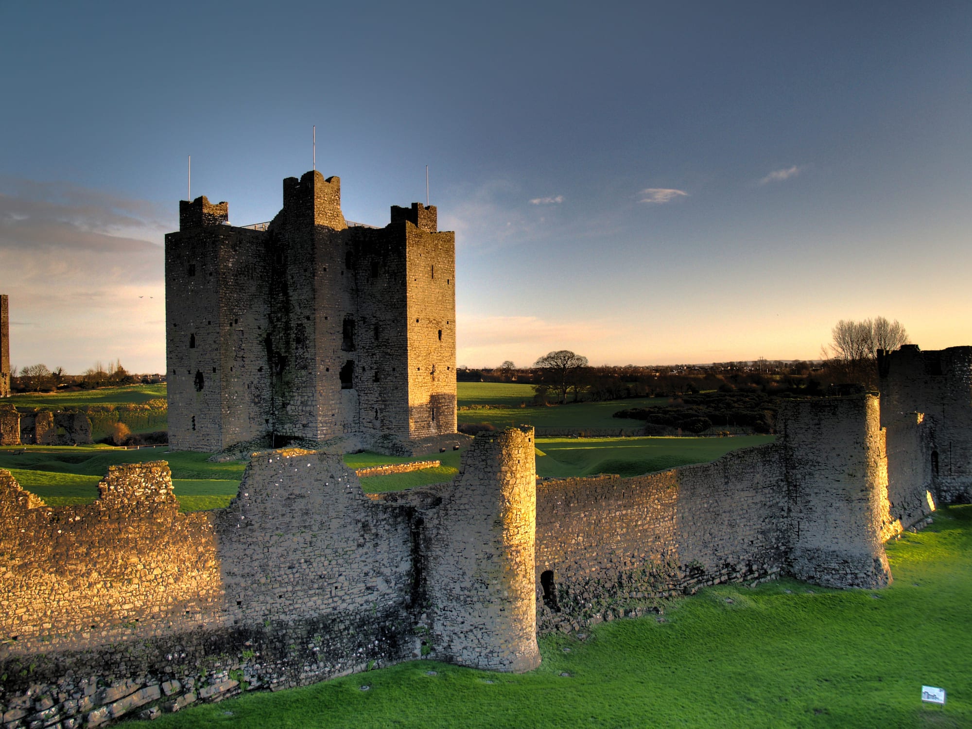 A tall square stone castle keep with corner towers, behind a ruined curtain wall with round turrets, lit by low sun