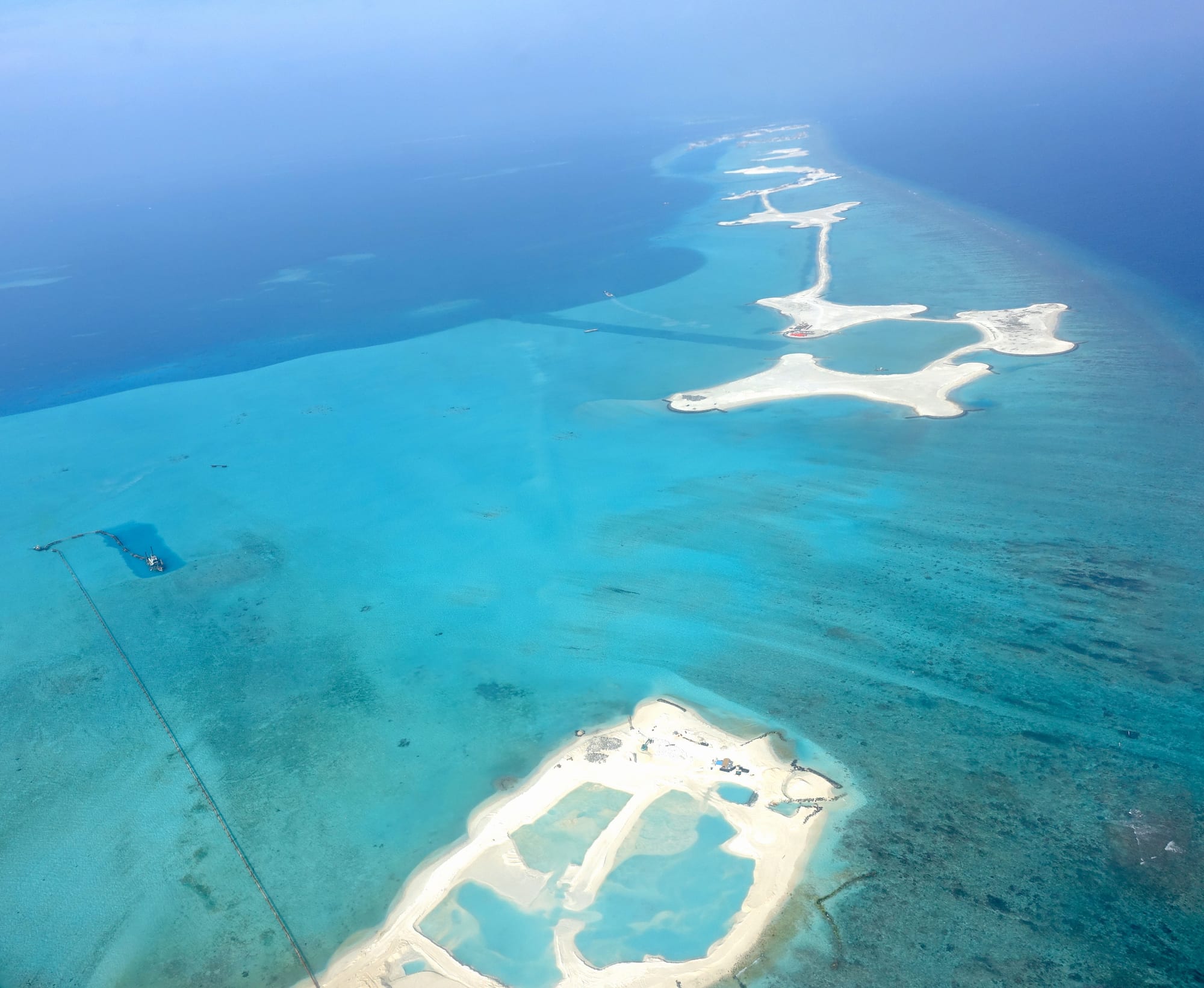 Bright white strips and blocks of newly pumped sand forming artificial islands on a pale turquoise reef flat, with a dredging pipeline