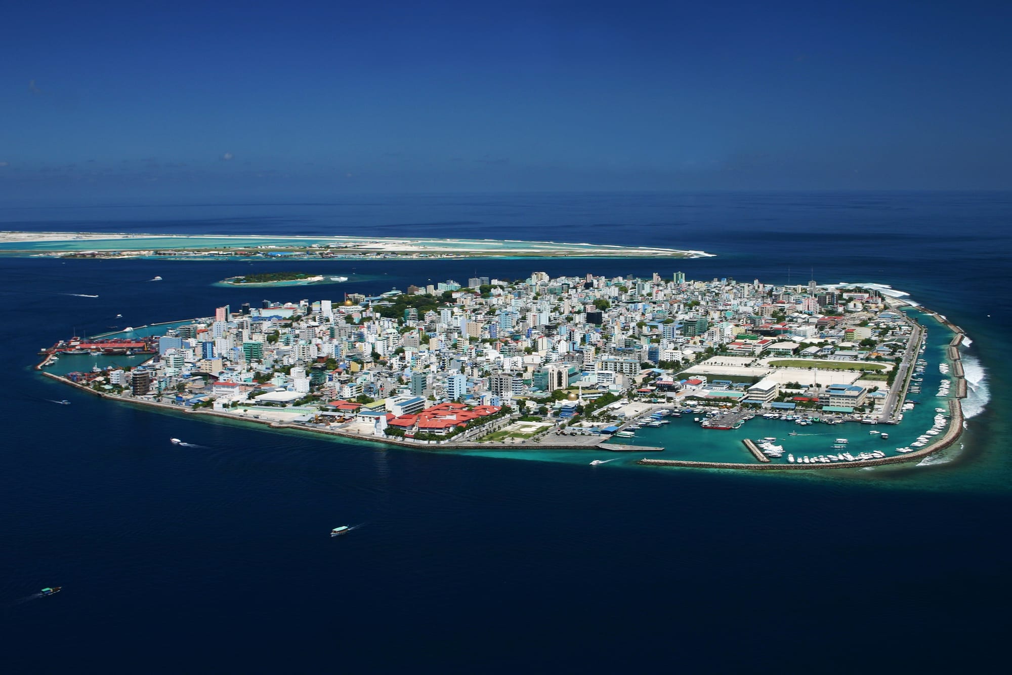 A small island completely covered with tightly packed buildings, ringed by harbours and sea walls, with a long flat airport island behind