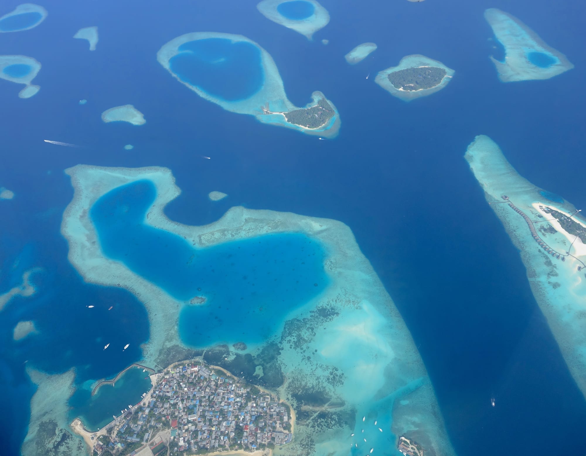 An aerial view of turquoise coral reefs and blue lagoons scattered across deep blue sea, with a densely built island at the bottom
