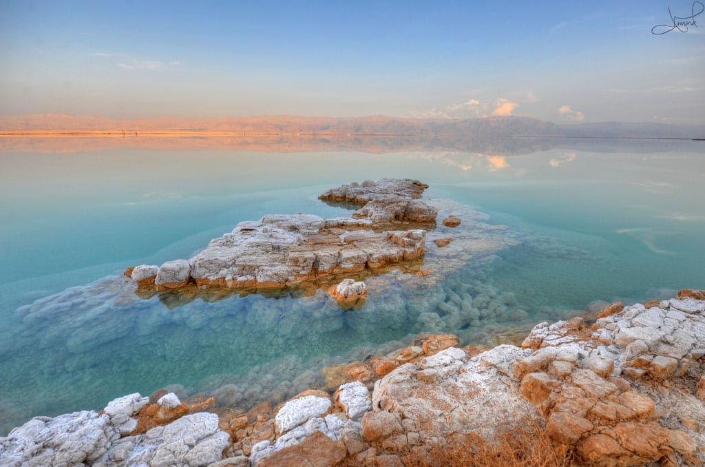 A still, pale-blue salt lake with white mineral crusts along the shore and bare mountains behind