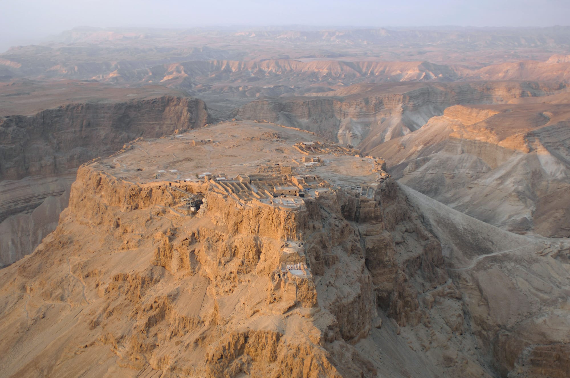 A flat-topped desert mesa ringed by cliffs, ruined buildings on top, the Dead Sea beyond