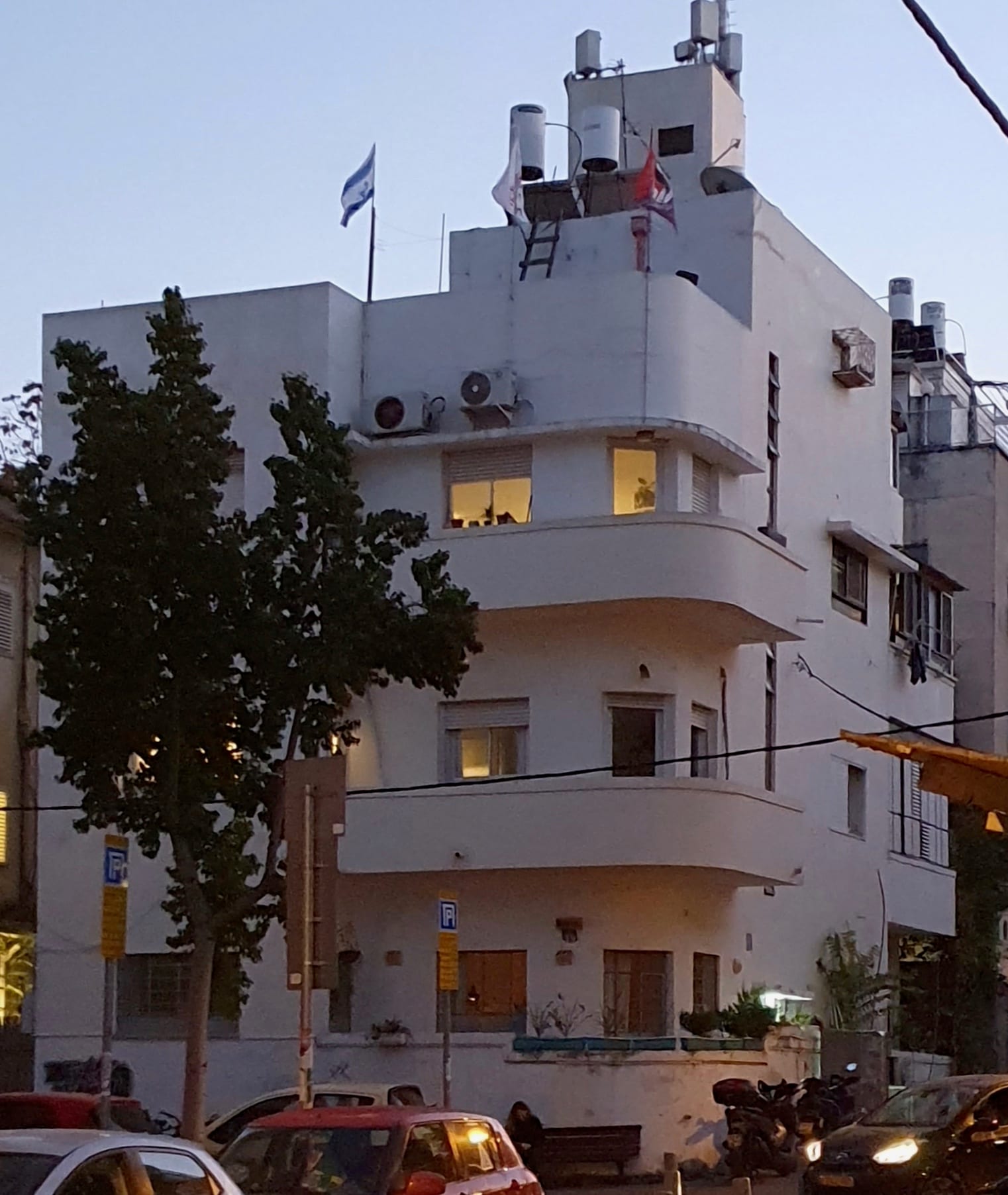A white, curved-balcony apartment building at dusk with lit windows, air-conditioning units and a rooftop water heater